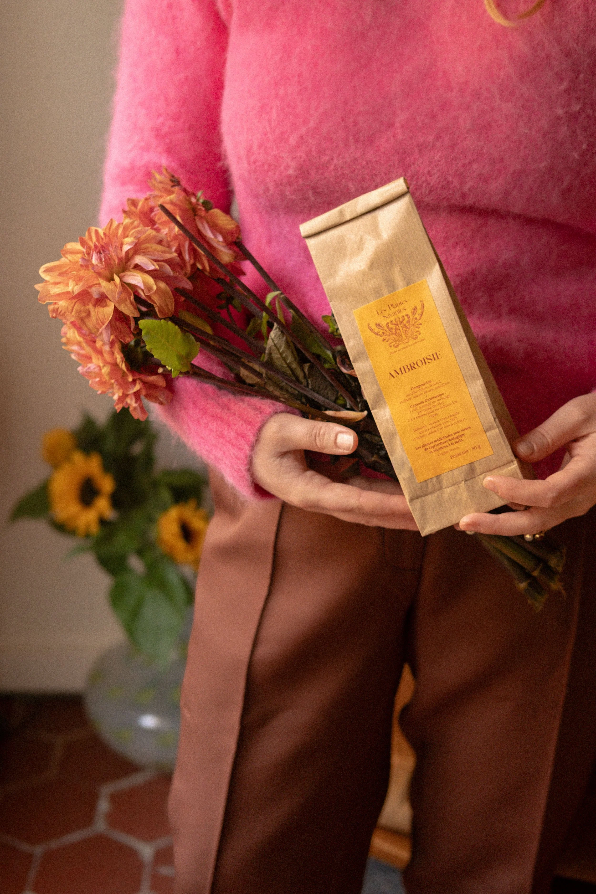 Une personne tient un bouquet de fleurs oranges et une boîte de thé en papier marron avec une étiquette jaune. Un vase avec des tournesols est visible en arrière-plan.