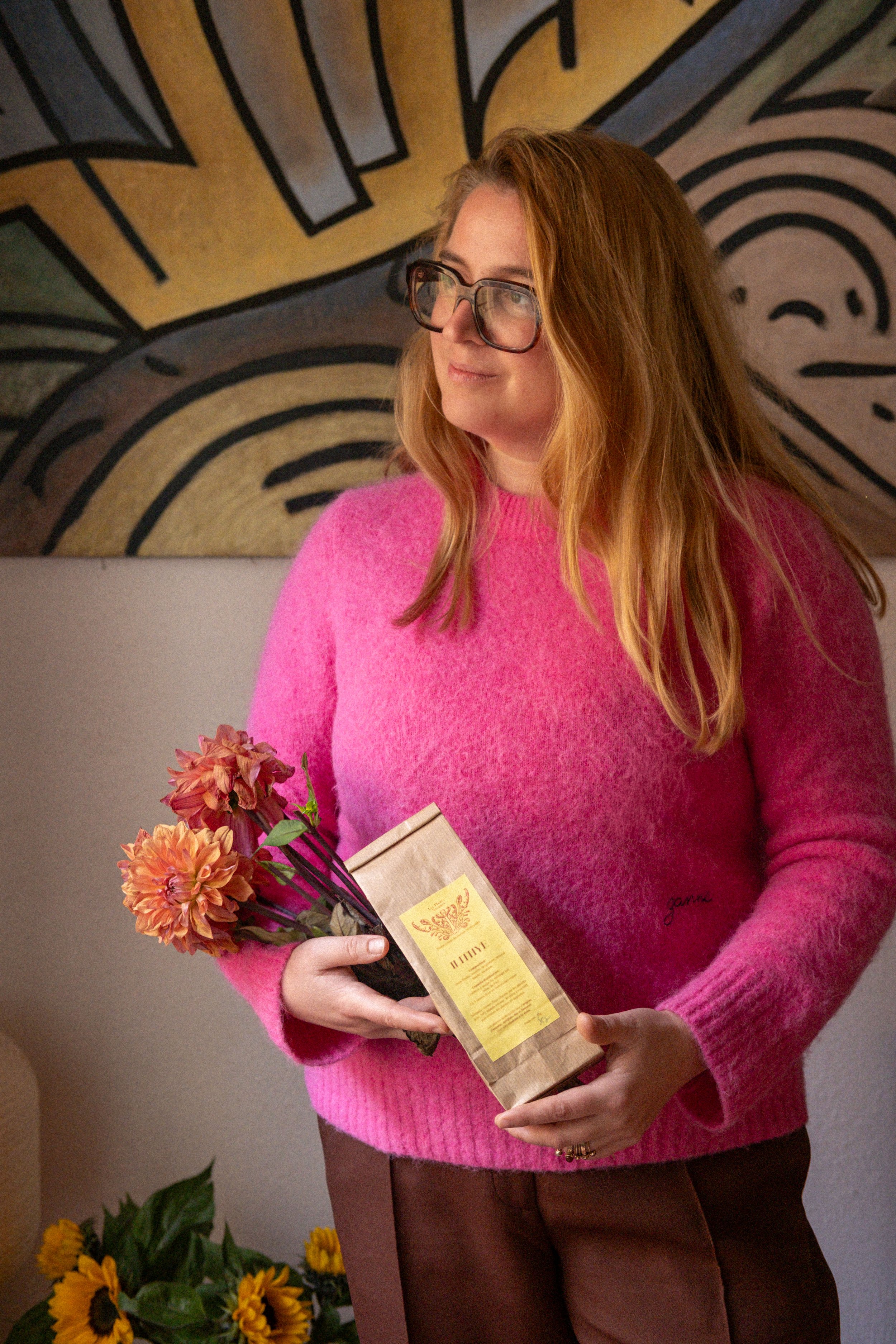 Une femme aux cheveux roux portant des lunettes et un pull rose tient un bouquet de fleurs et une boîte de café dans une pièce décorée avec une œuvre d'art abstraite au mur.