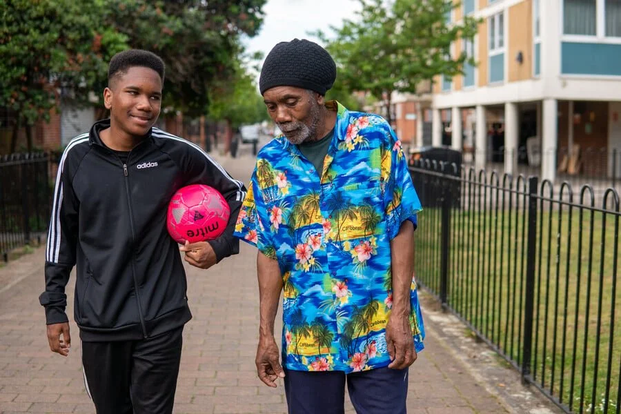 A young man holding a pink soccer ball walking beside an older man wearing a colorful Hawaiian shirt, both on a sidewalk near a black metal fence, with trees and apartment buildings in the background.