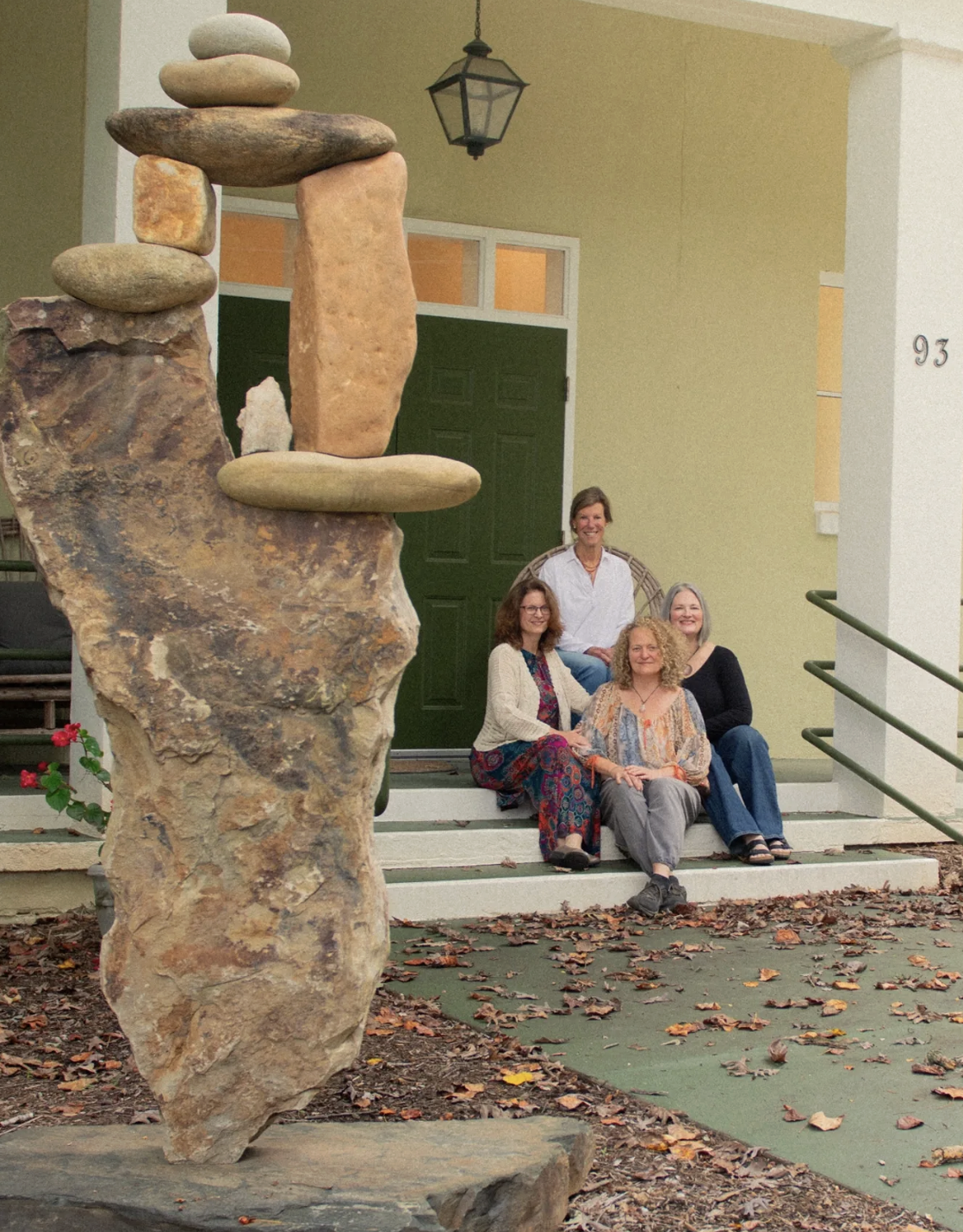 Four women sitting on front steps of a house, smiling, with a large stone sculpture in the foreground.