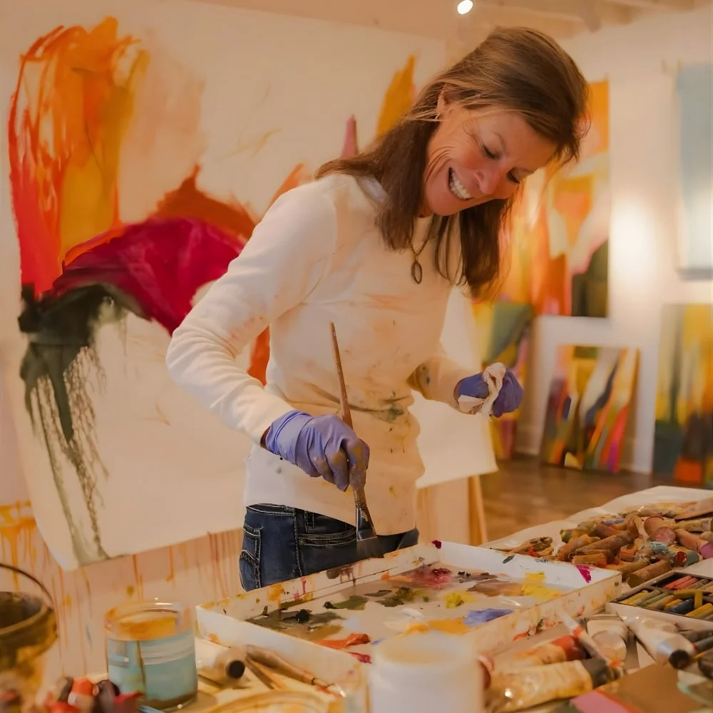 Caroline Young is smiling while painting on a canvas in an art studio, surrounded by colorful artwork and art supplies.