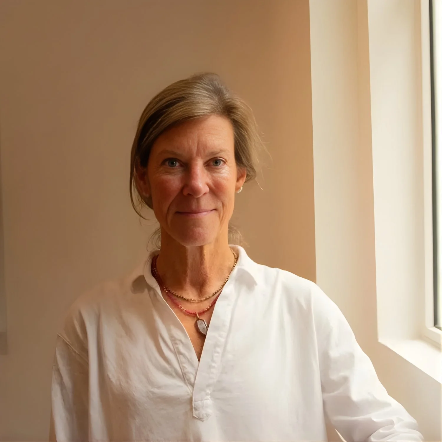 Portrait of a middle-aged woman with short light brown hair, wearing a white shirt and layered necklaces, standing near a window with beige walls in the background.