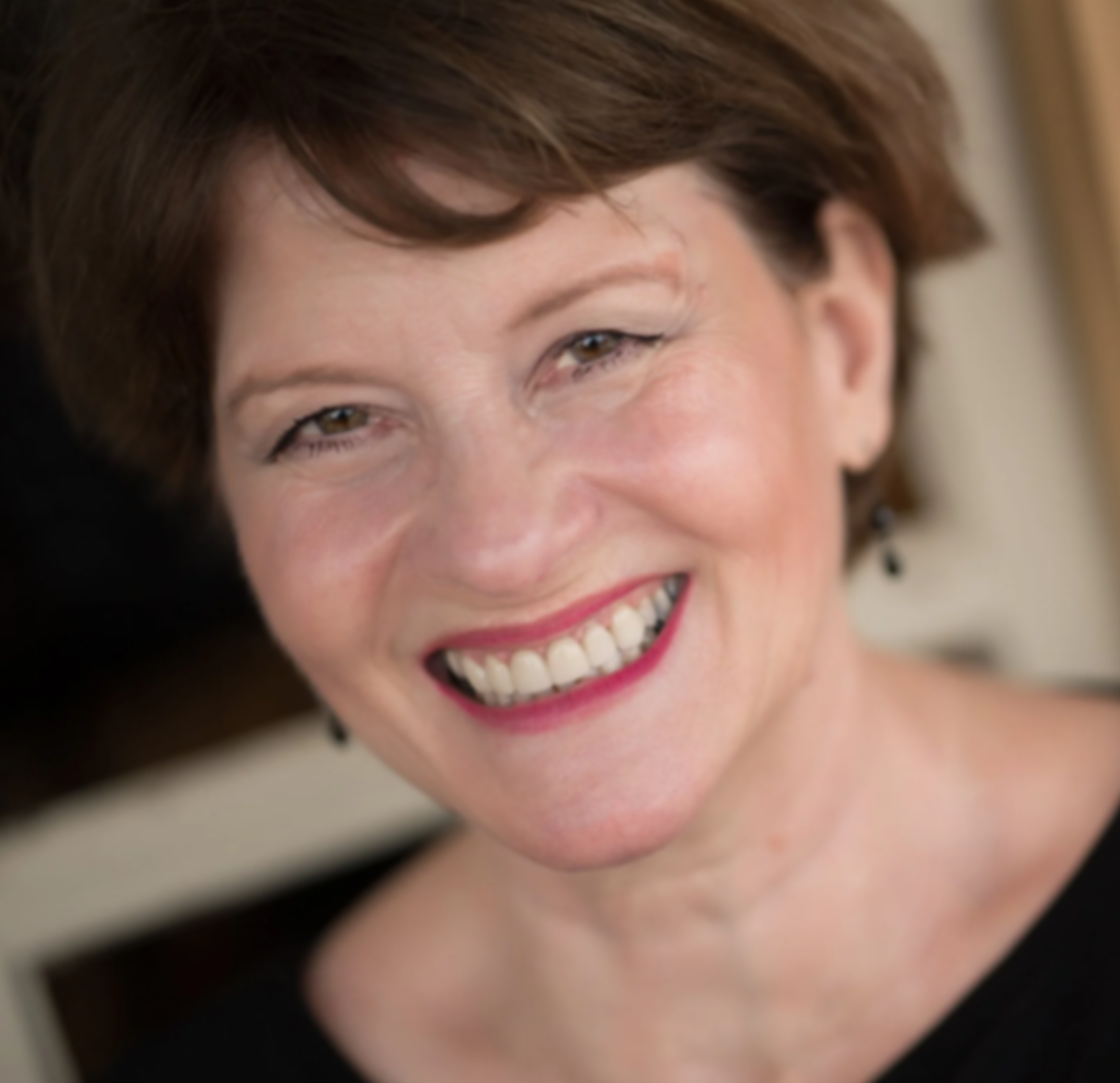 A woman with short brown hair smiling brightly, showing her teeth, wearing black earrings and a black top.
