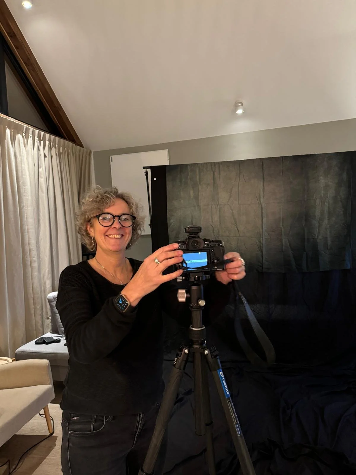 A woman with curly gray hair, glasses, and a black shirt is standing and smiling while adjusting a camera on a tripod in a living room.