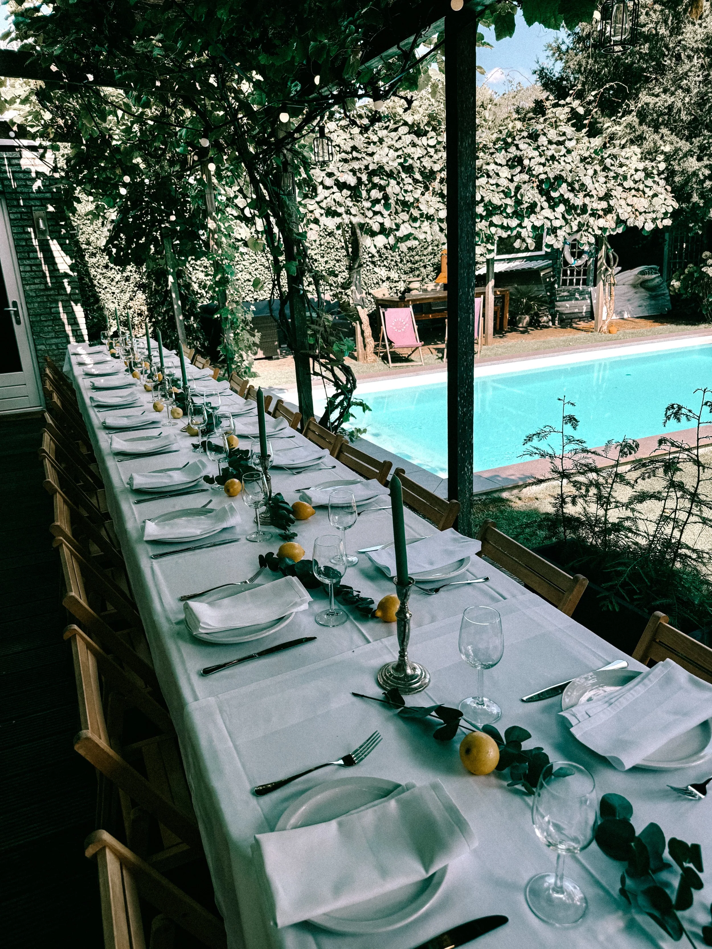 Long outdoor dining table set with white plates, silverware, wine glasses, white napkins, green candles, and yellow lemons, arranged on a white tablecloth under a leafy canopy near a swimming pool.