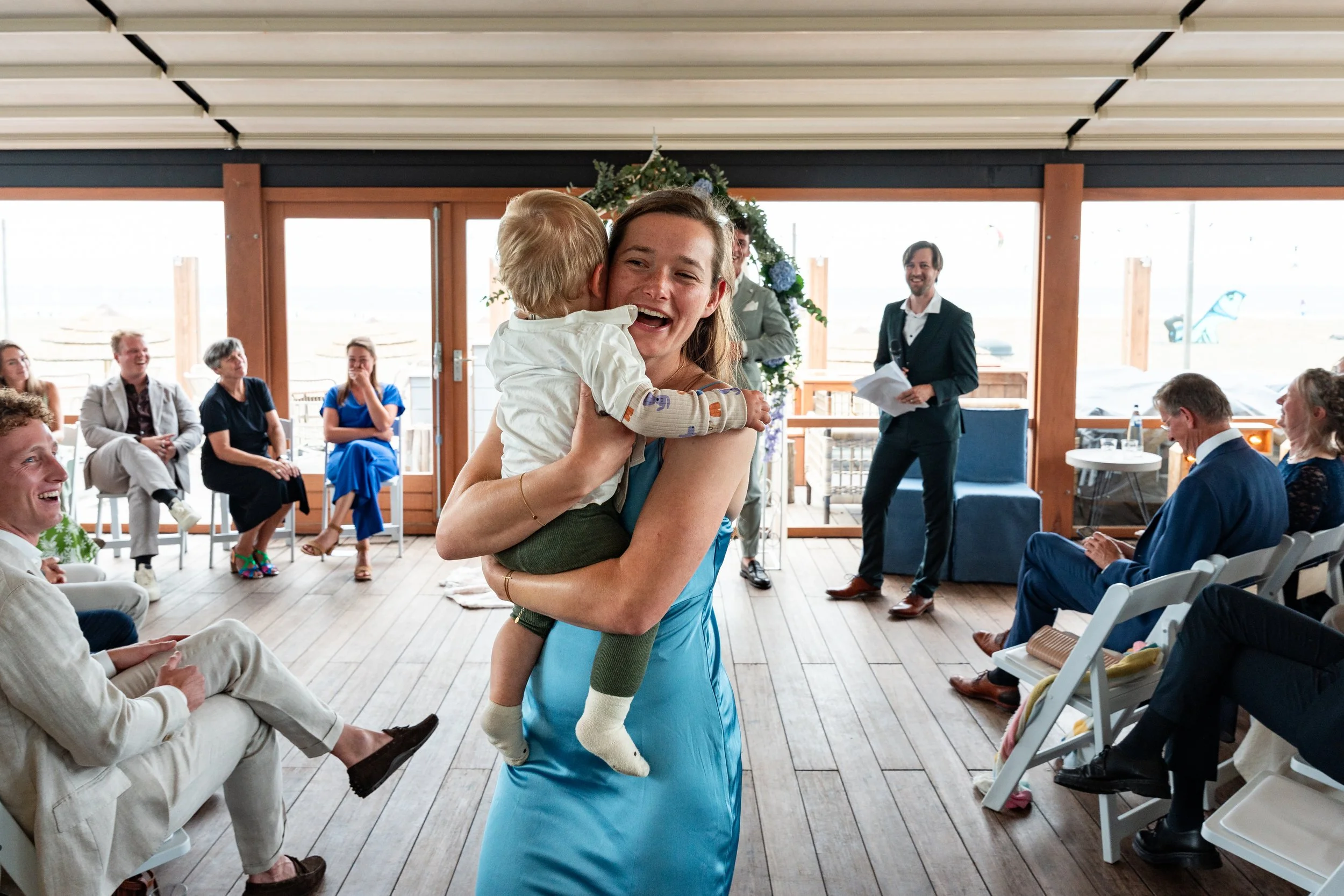 A woman holding a young boy and smiling inside a decorated room during a celebration, with several seated guests and a man with a microphone in the background.
