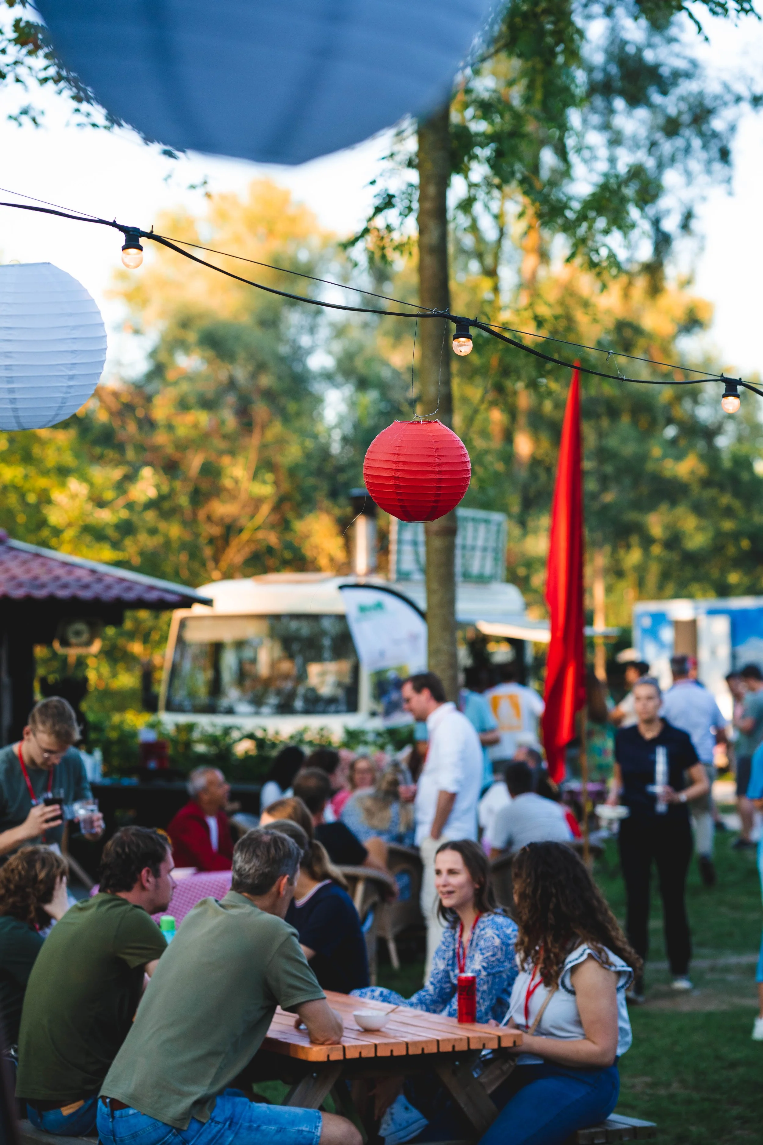 People gathered at an outdoor event with string lights and paper lanterns hanging overhead, with food trucks and trees in the background.
