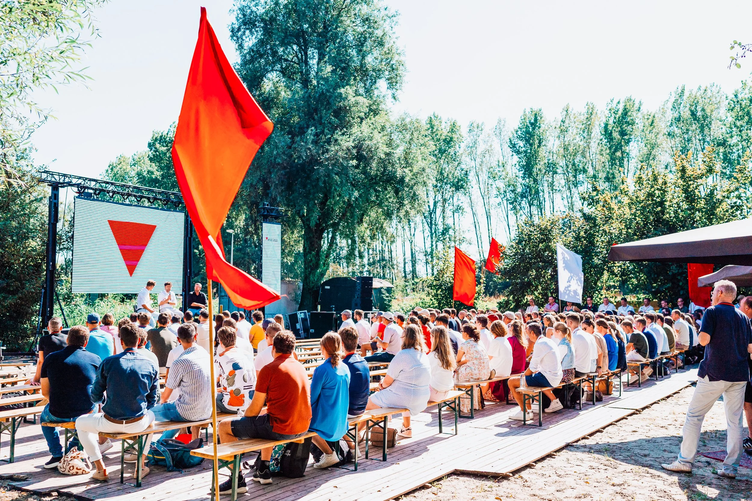 Outdoor event with a large crowd seated on benches, facing a stage with a large screen displaying a red triangle. There are several red flags and a few white flags, surrounded by trees and sunny weather.