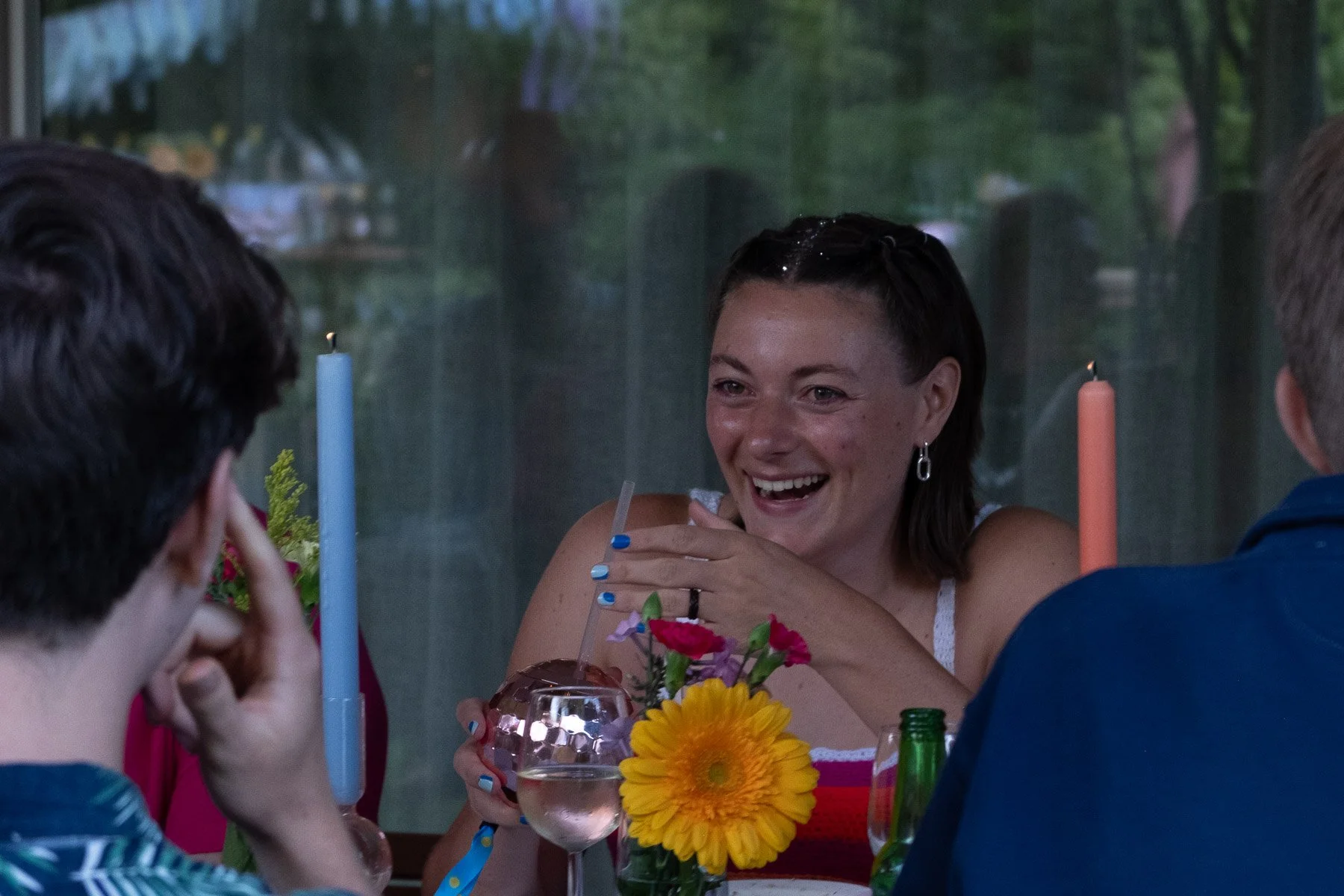 A woman laughing and celebrating at a dinner table with friends, holding a drink with a straw, with flowers and candles on the table, in a cozy indoor setting.