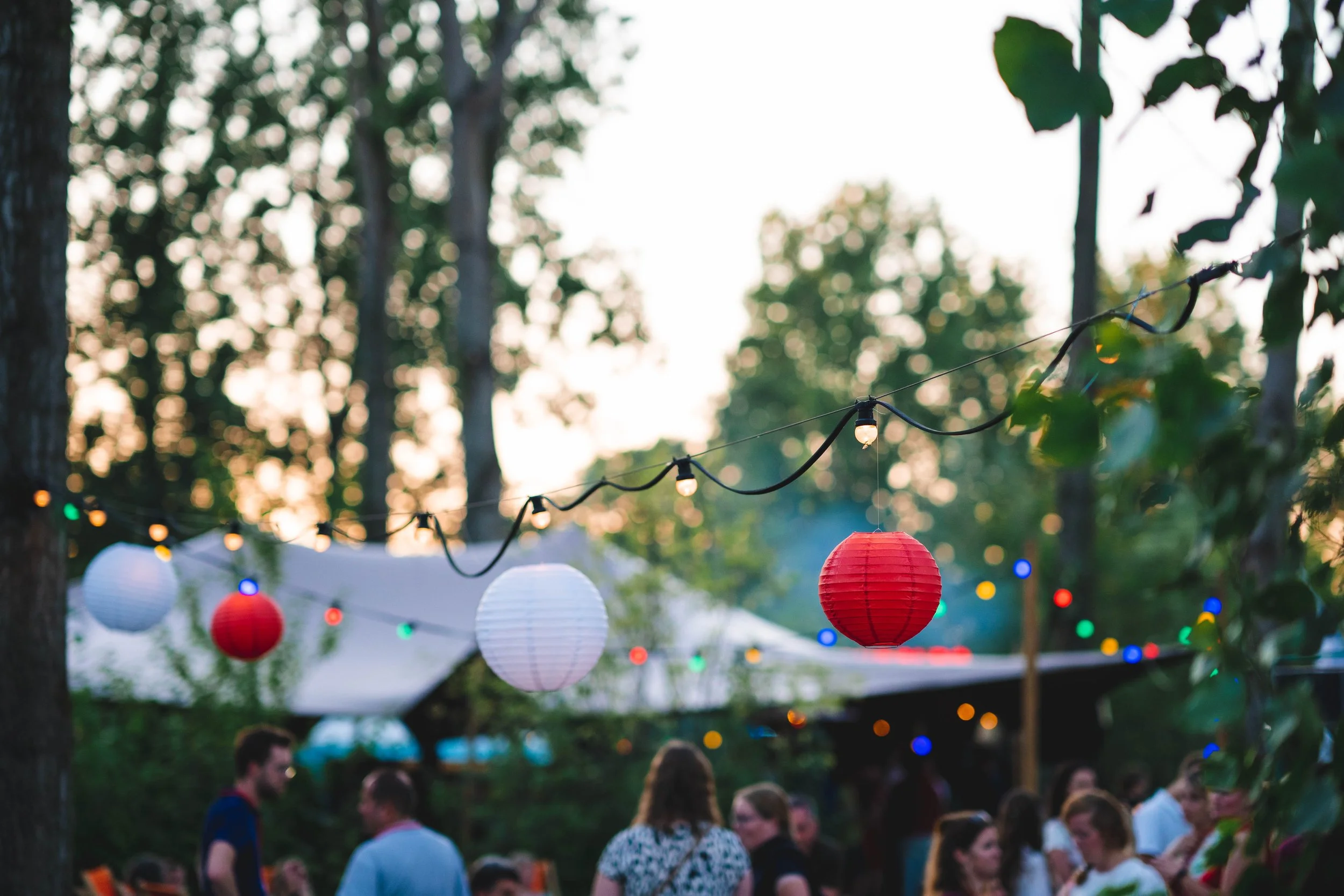 Outdoor gathering with string lights and paper lanterns hanging among trees, with people socializing in the background during the evening.