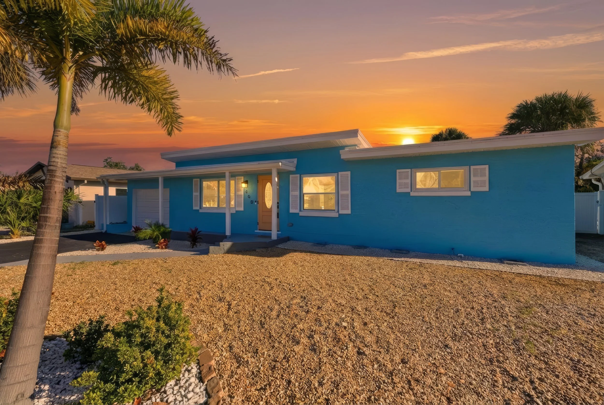 A single-story blue house during sunset with a front porch, white shutters, and decorative plants in the yard, including a palm tree in the foreground.