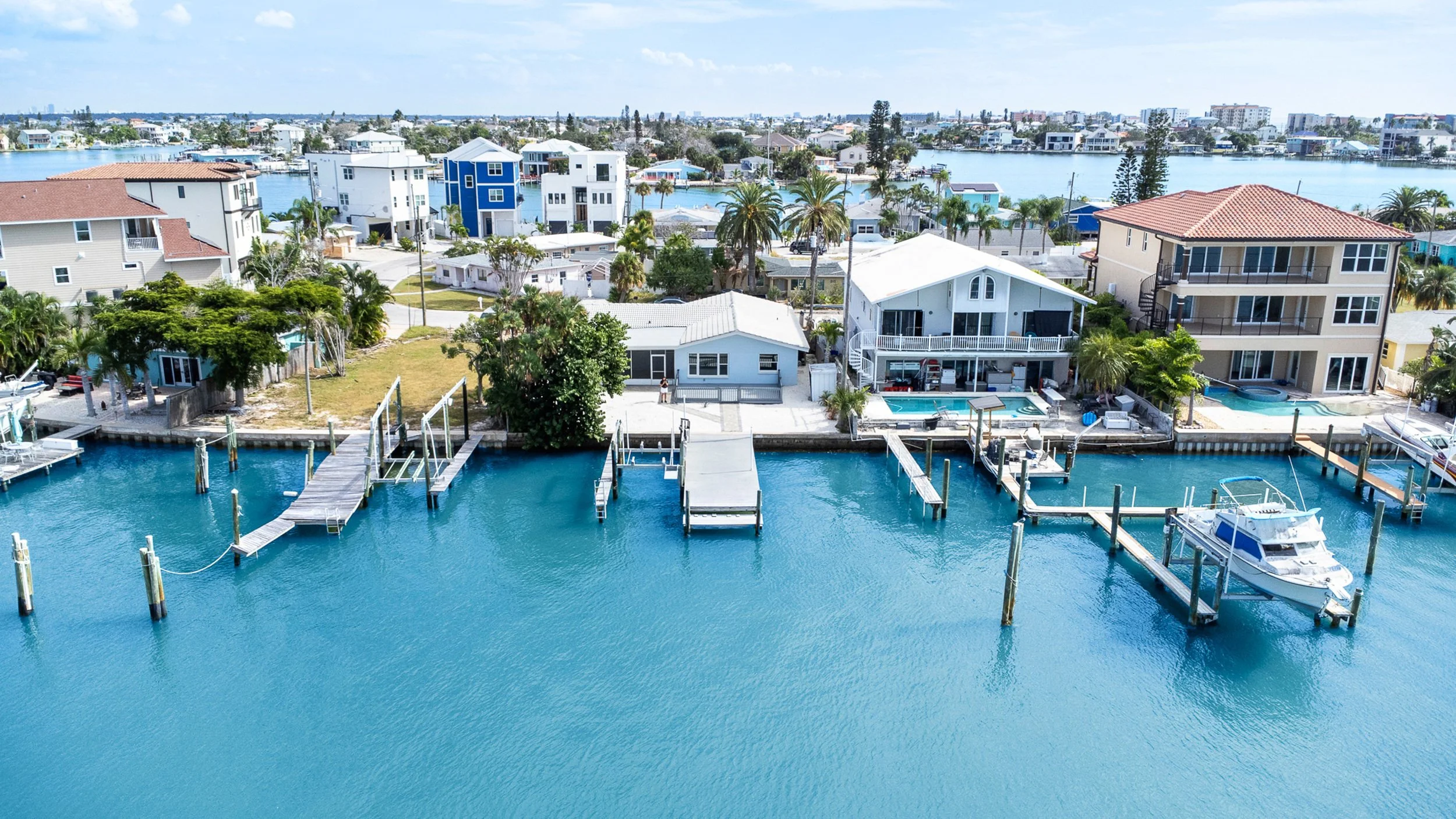 Aerial view of waterfront houses with docks and boats on a sunny day, with a canal and city skyline in the background.