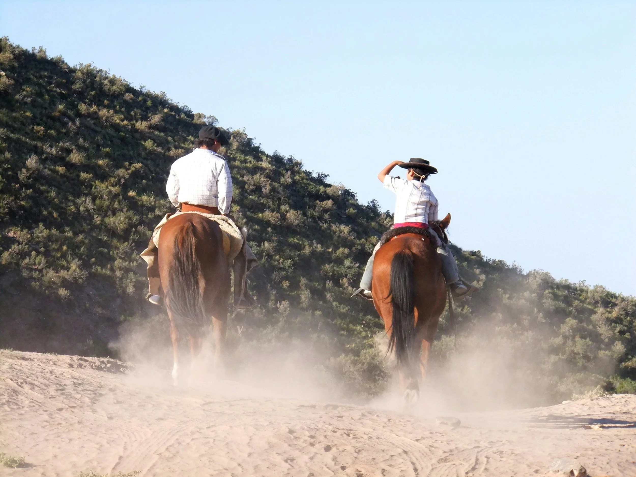 Two people riding horses on a dusty trail in a hilly, brush-covered landscape under a clear blue sky.