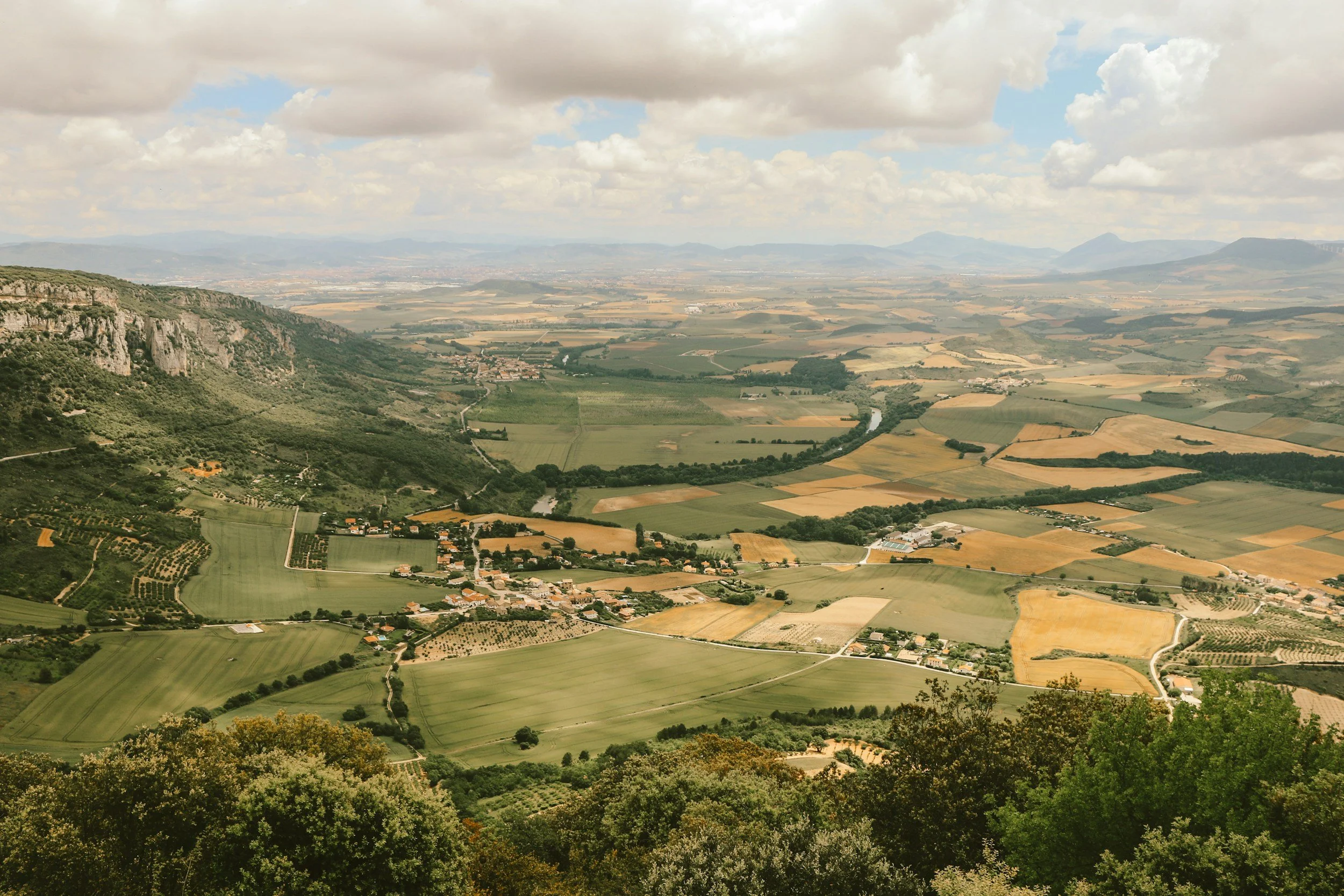A panoramic view of a rural landscape with fields, small villages, winding roads, a river, green hills, and mountains in the distance under a partly cloudy sky.