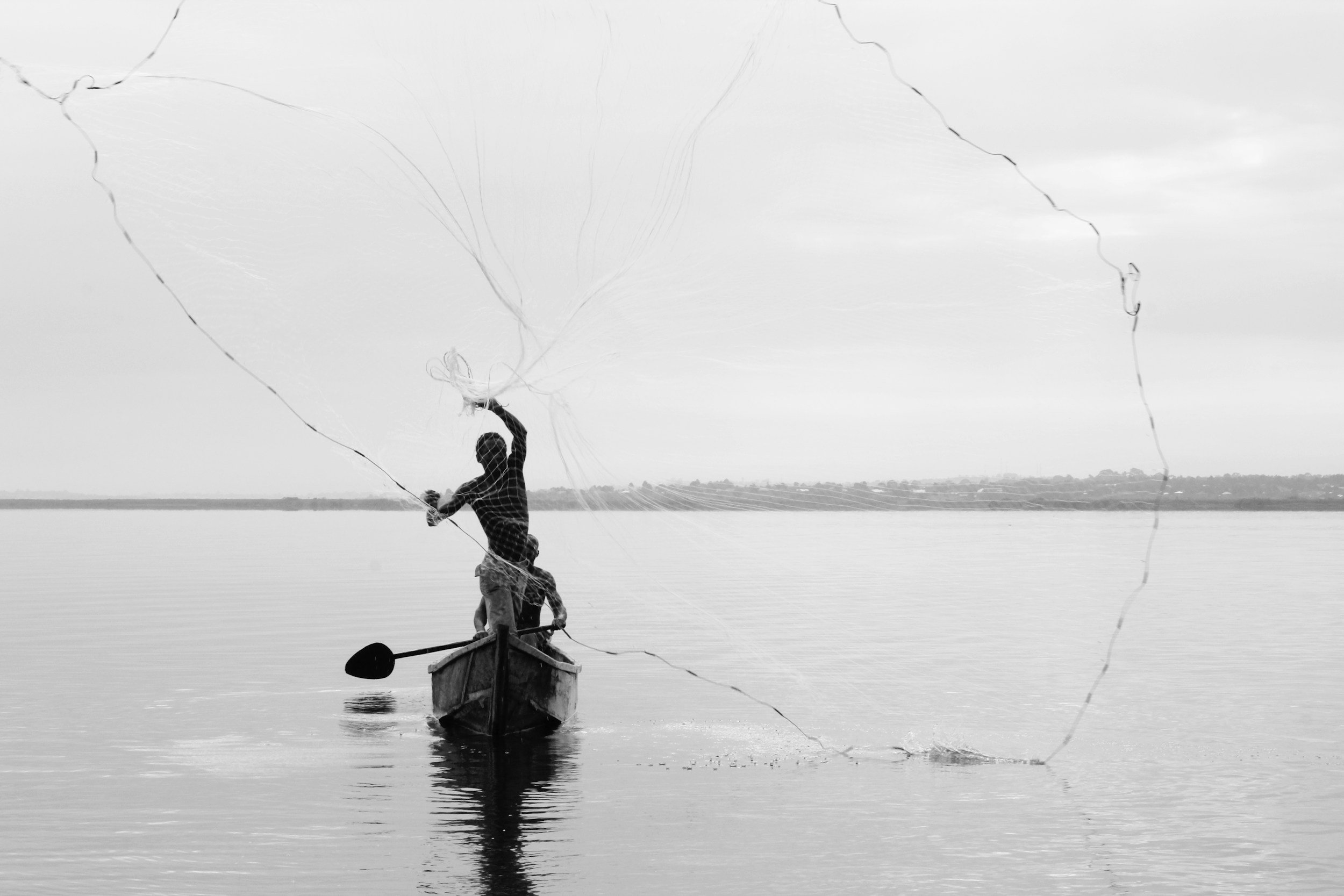 A person standing in a small boat on a calm body of water, casting a fishing net.