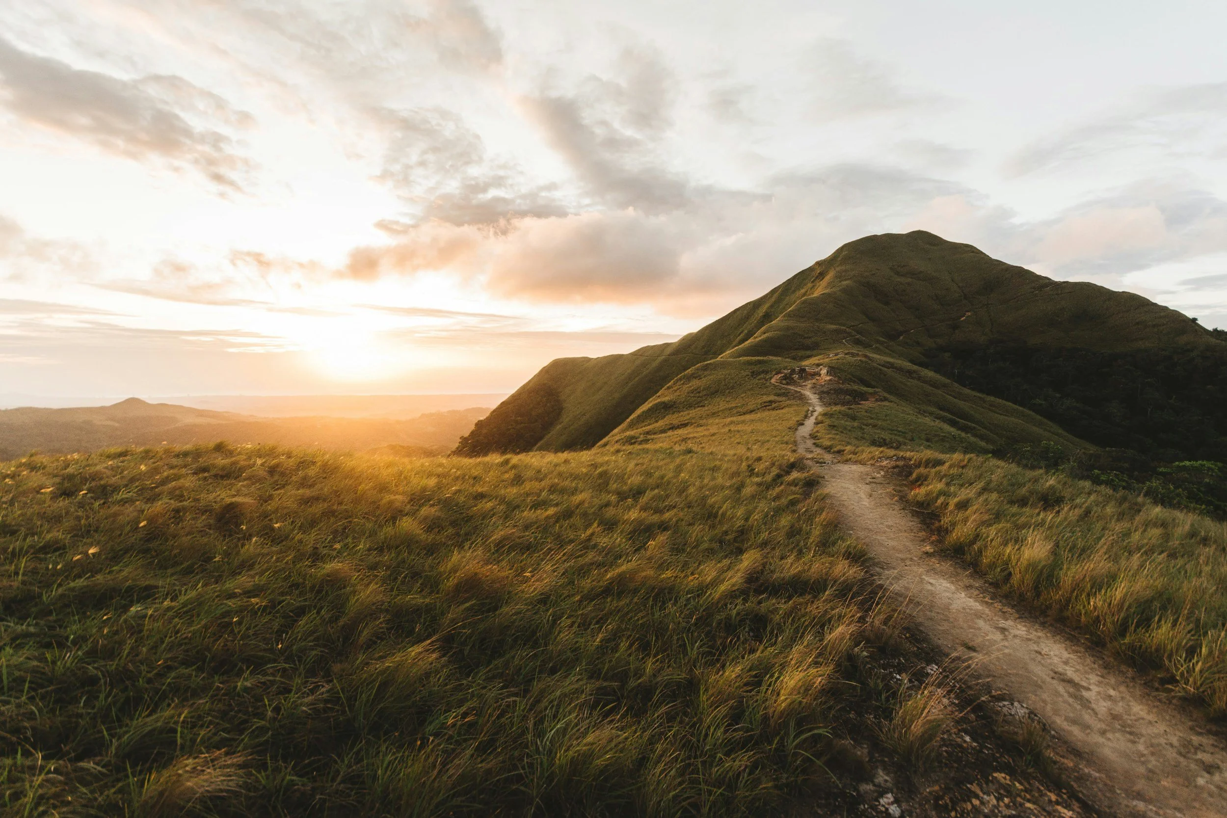 A hiking trail winding through grassy hills with a mountain in the background during sunset.
