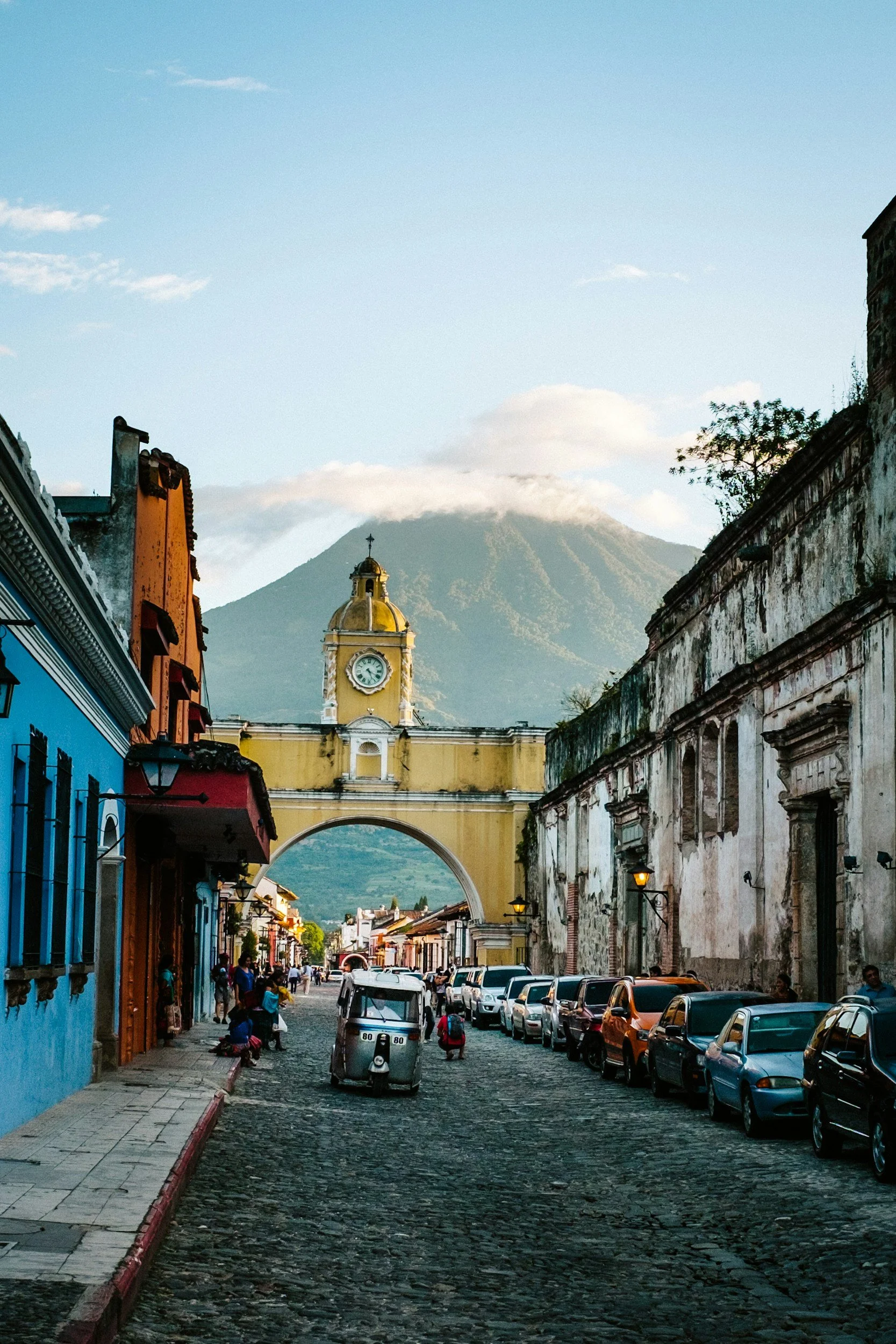 A cobblestone street in Antigua, Guatemala, with colorful colonial buildings, parked cars, and a yellow church archway with a clock, volcano in the background.