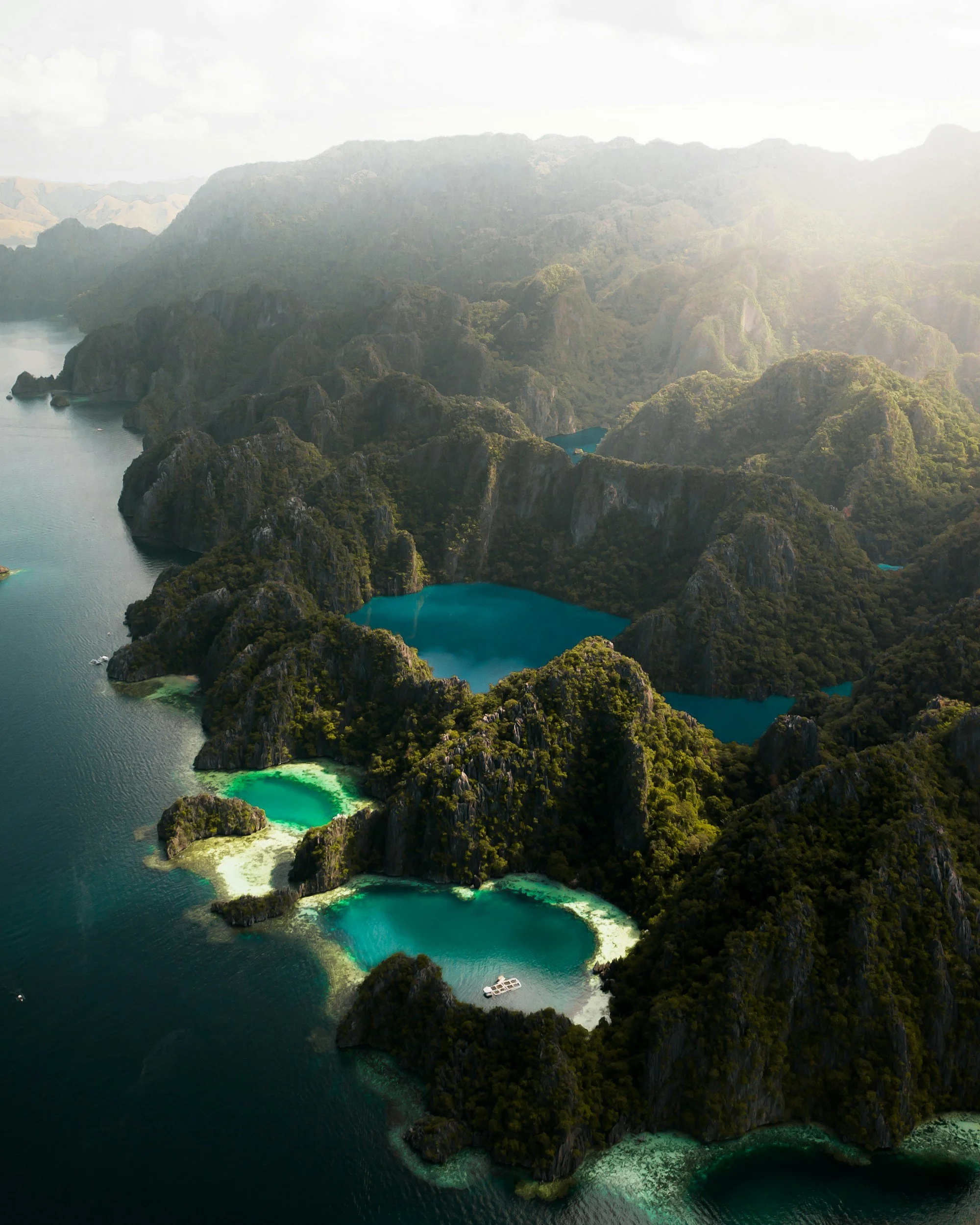 Aerial view of a tropical archipelago with several turquoise lakes among green forested limestone cliffs surrounded by dark blue ocean.