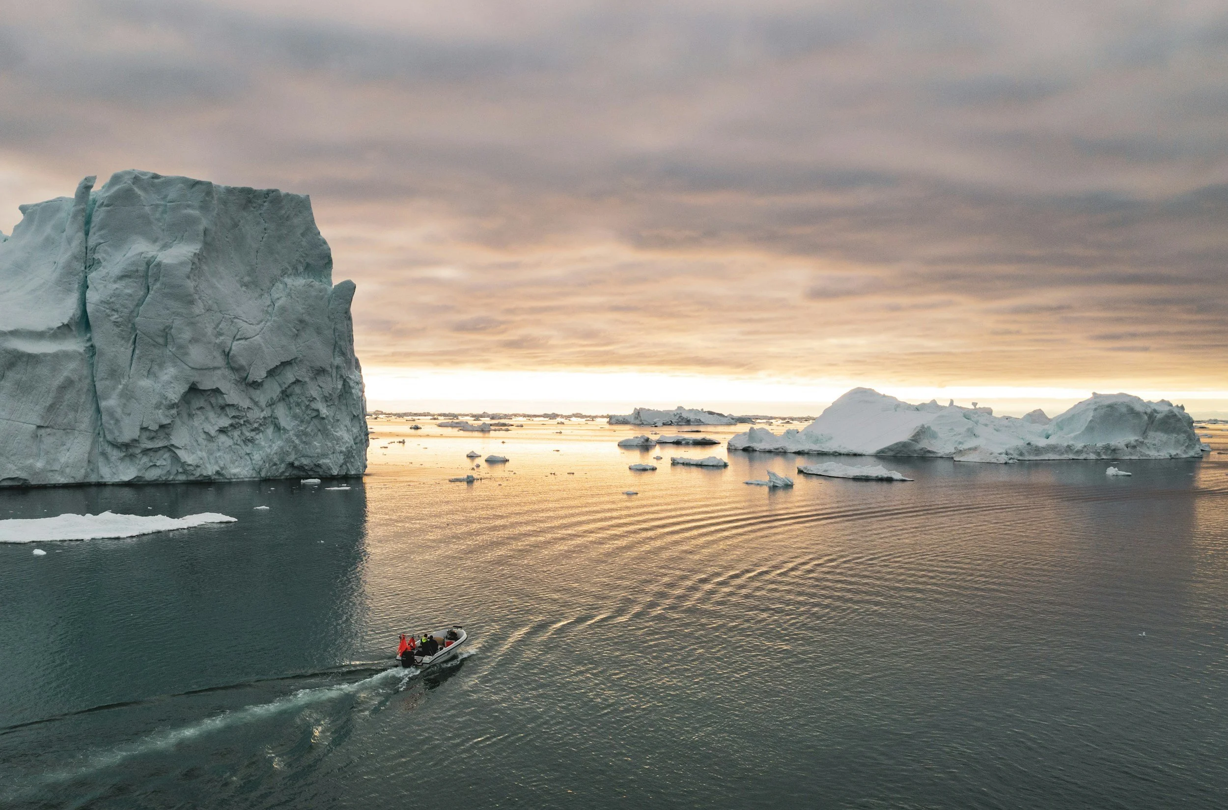 A boat moving through icy water near large icebergs and floating ice in a polar region during sunset or sunrise.