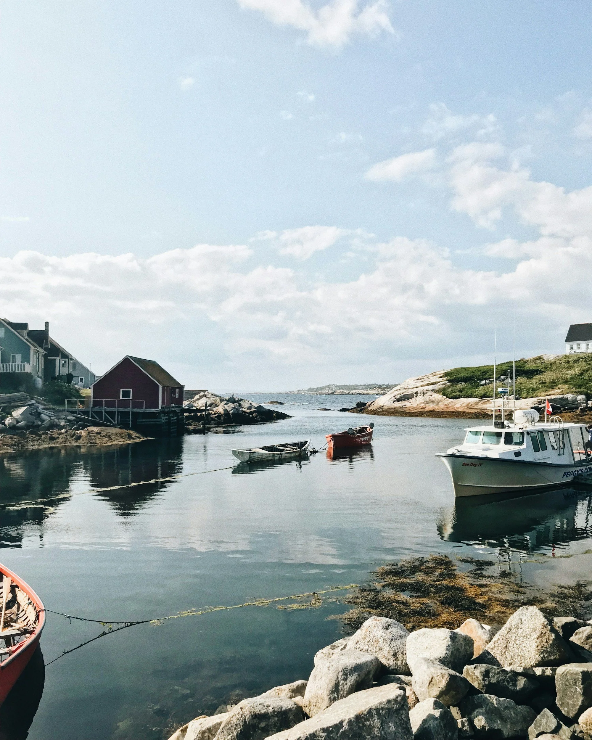A serene harbor scene with small boats docked near colorful houses on rocky shores under a partly cloudy sky.