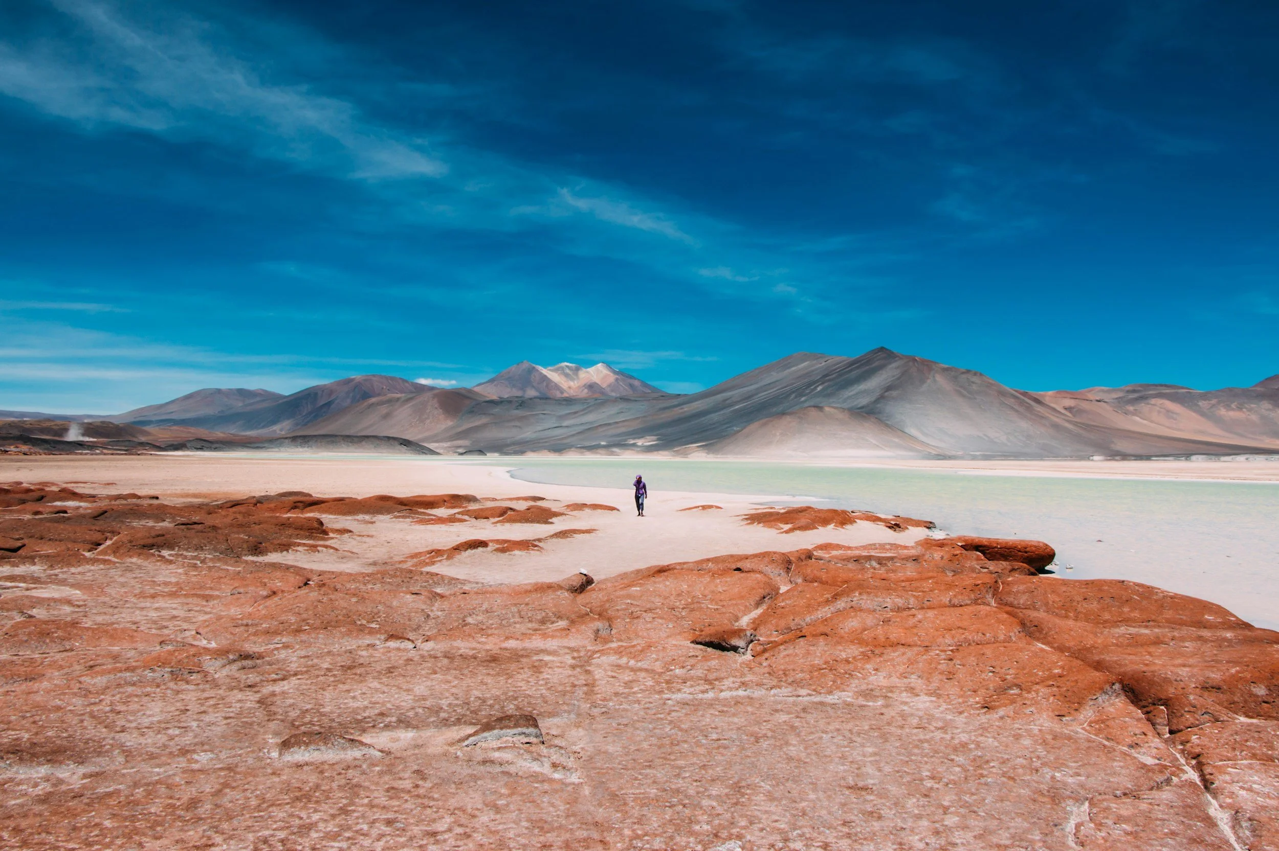 A person walking across a vast desert landscape with reddish rocks, a white salt flat, and mountains in the background under a blue sky.