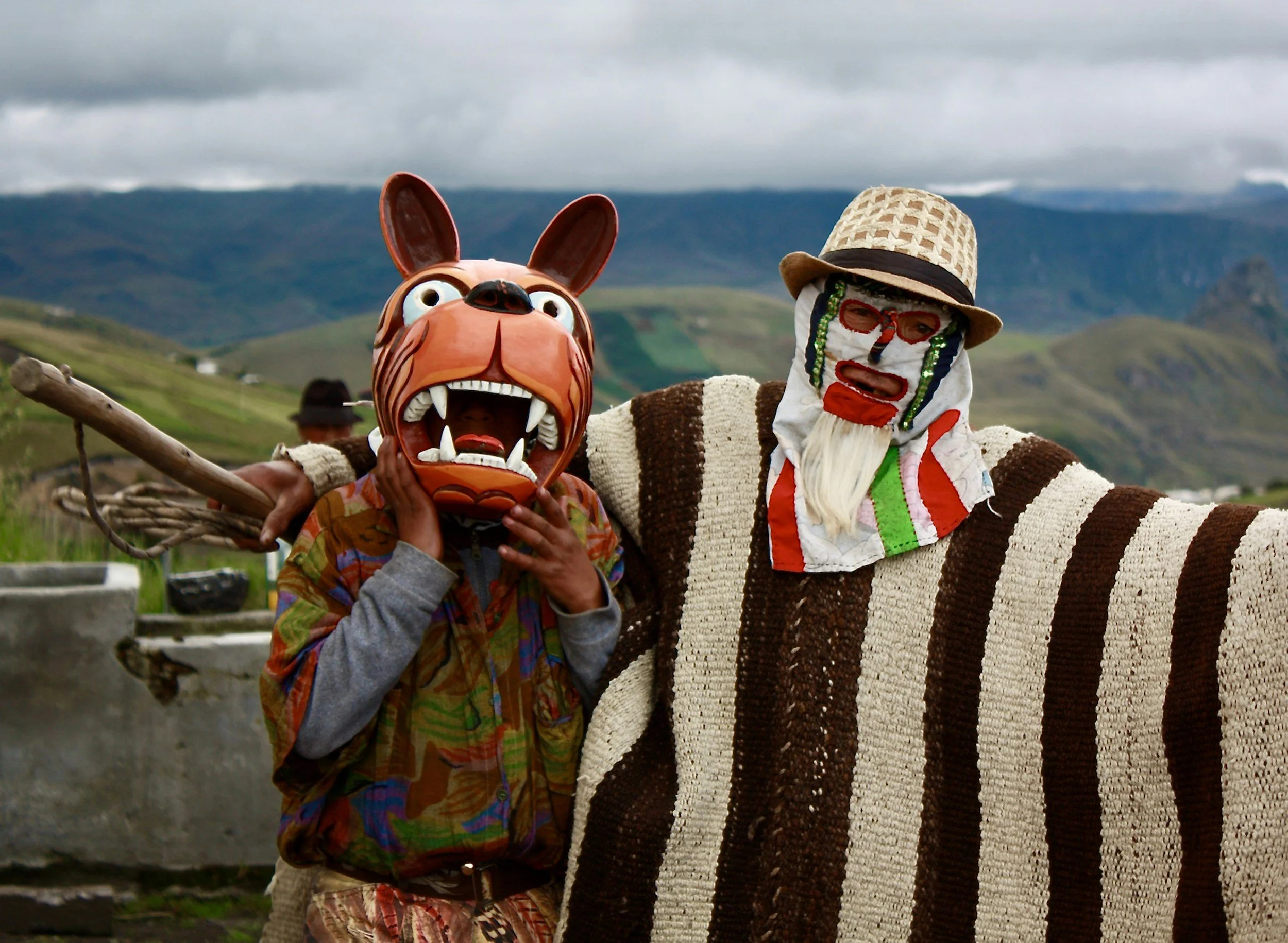 People in colorful costumes and masks standing outdoors with a scenic mountain landscape in the background.