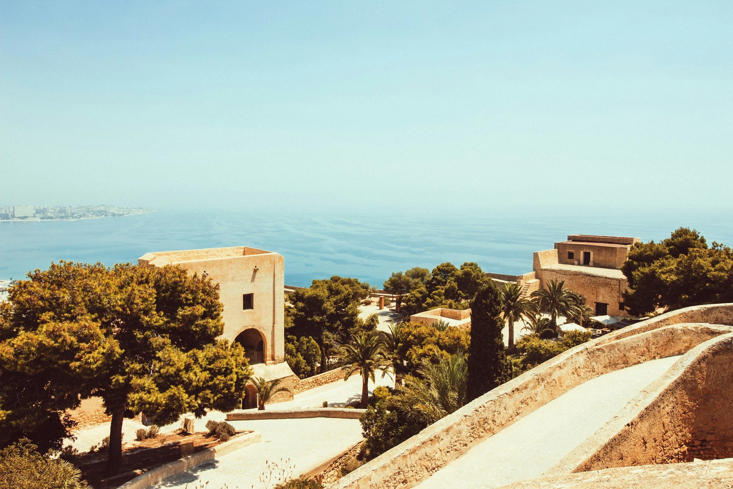 A scenic view of historic stone buildings and lush greenery on a hillside overlooking the ocean, with a distant shoreline visible on the horizon.