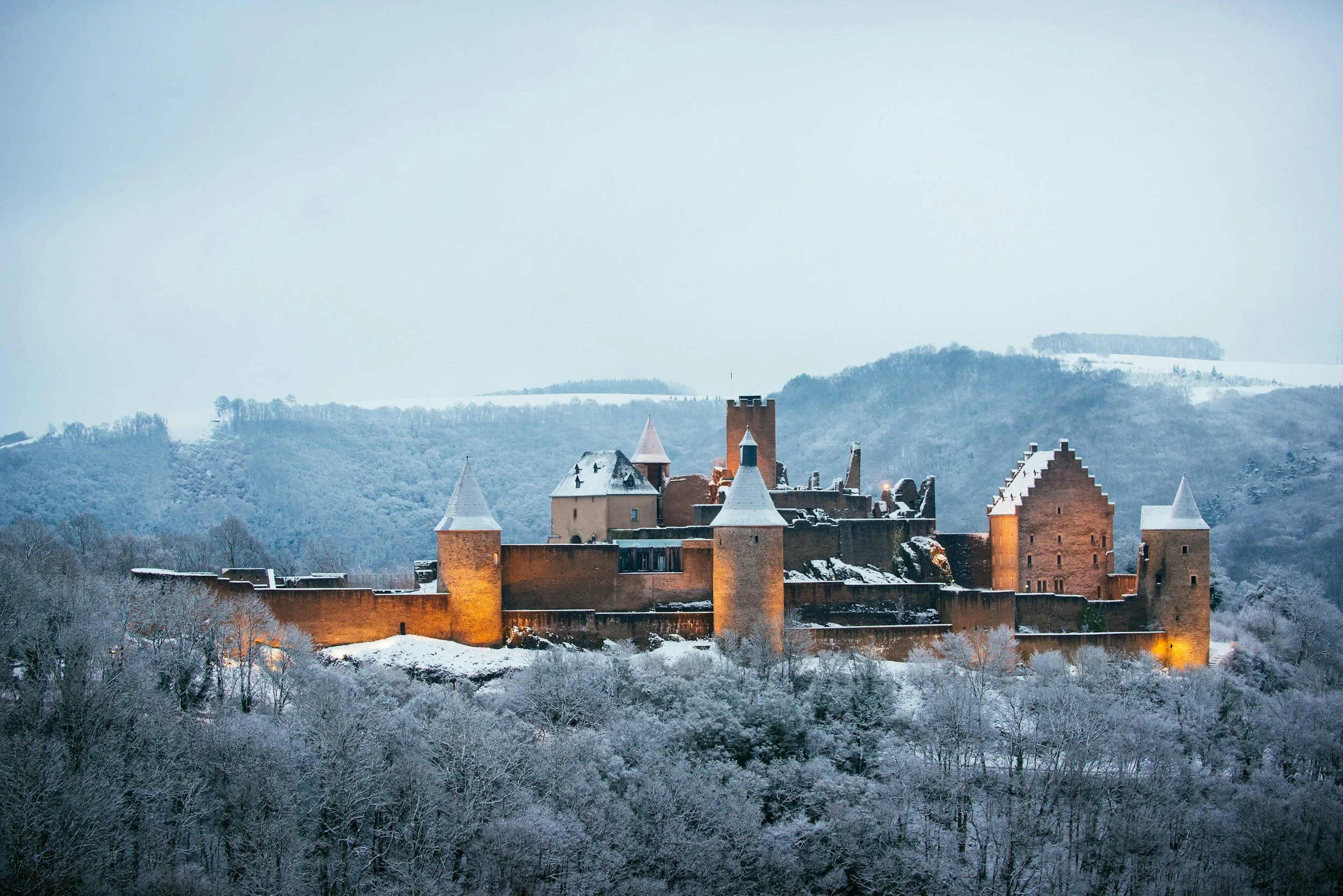 Snow-covered medieval castle on a hilltop with lit towers surrounded by snow-laden trees and mountains in the background.