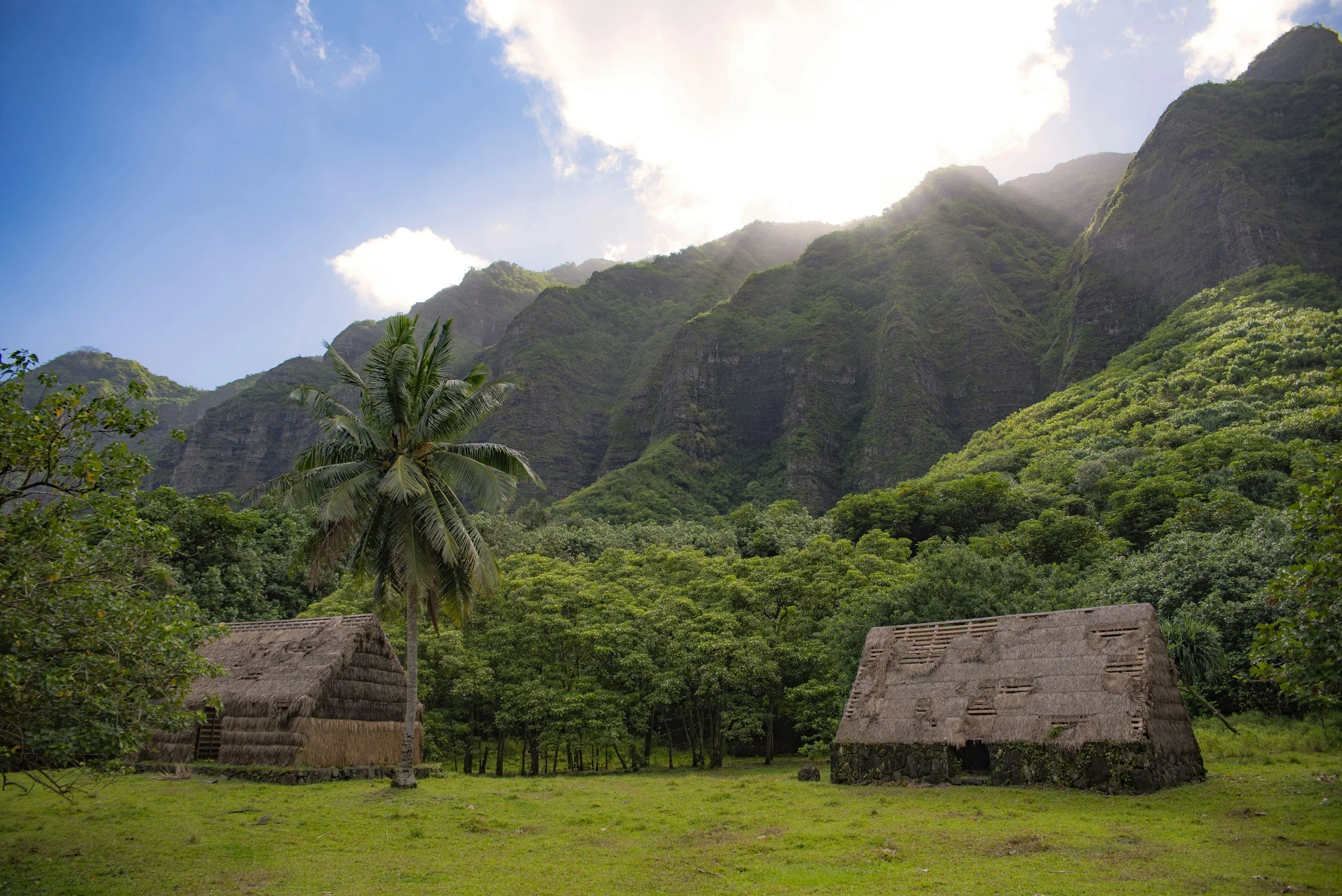 Two traditional thatched-roof huts are in a grassy clearing surrounded by lush green trees and dense forest. Behind the trees are tall, steep, green mountains with partially cloudy sky above.