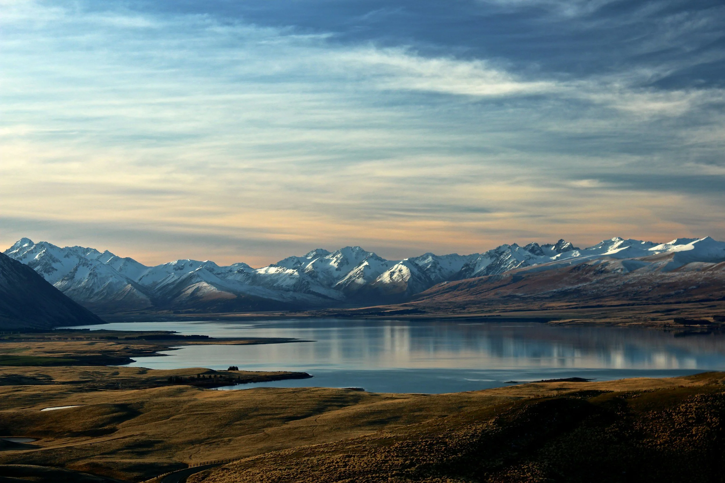 Scenic landscape of snow-capped mountains, a large calm lake reflecting the sky, and grassy plains in the foreground under a partly cloudy sky.