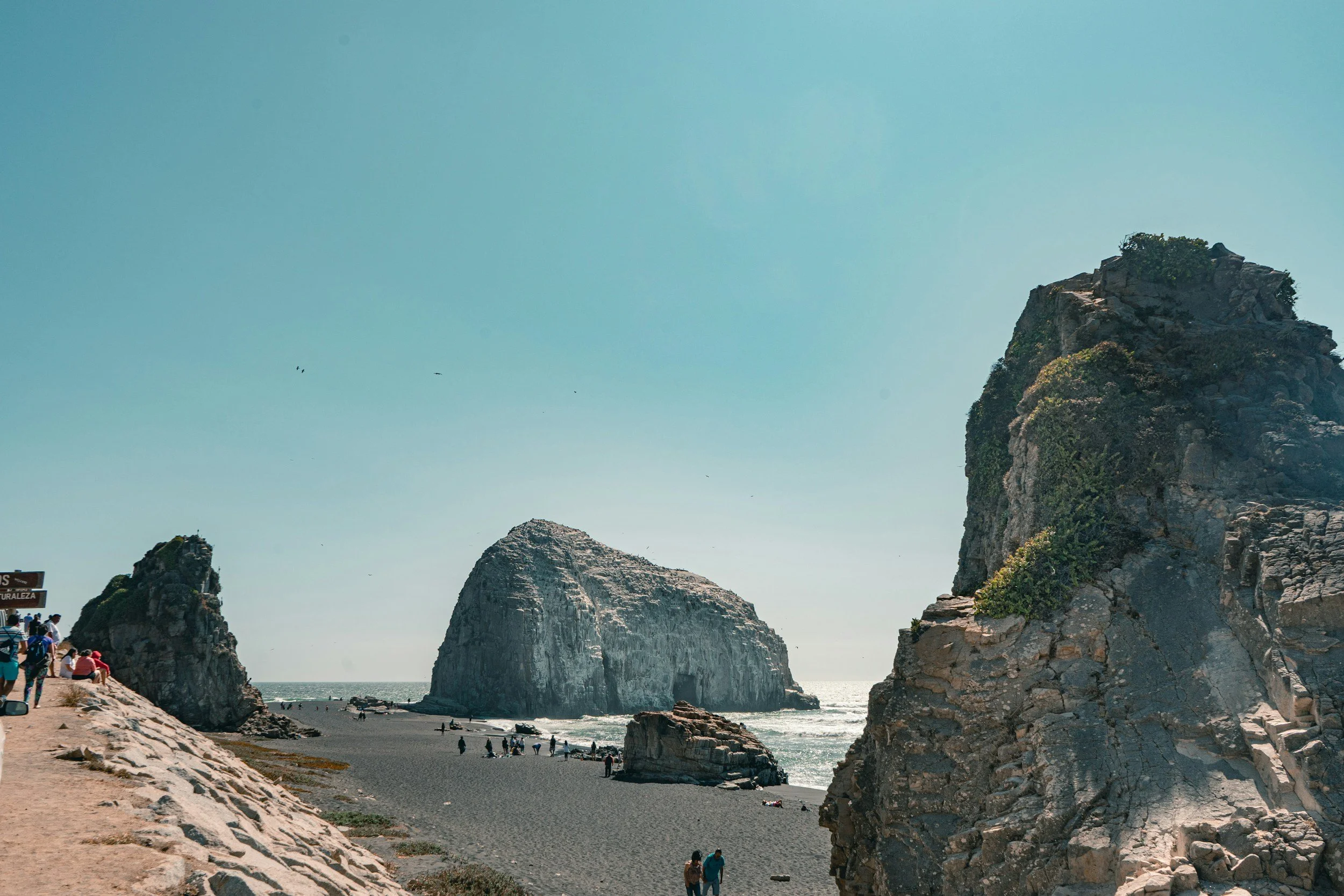 A coastal landscape with large rocky formations, a sandy beach, and people walking and relaxing near the water.