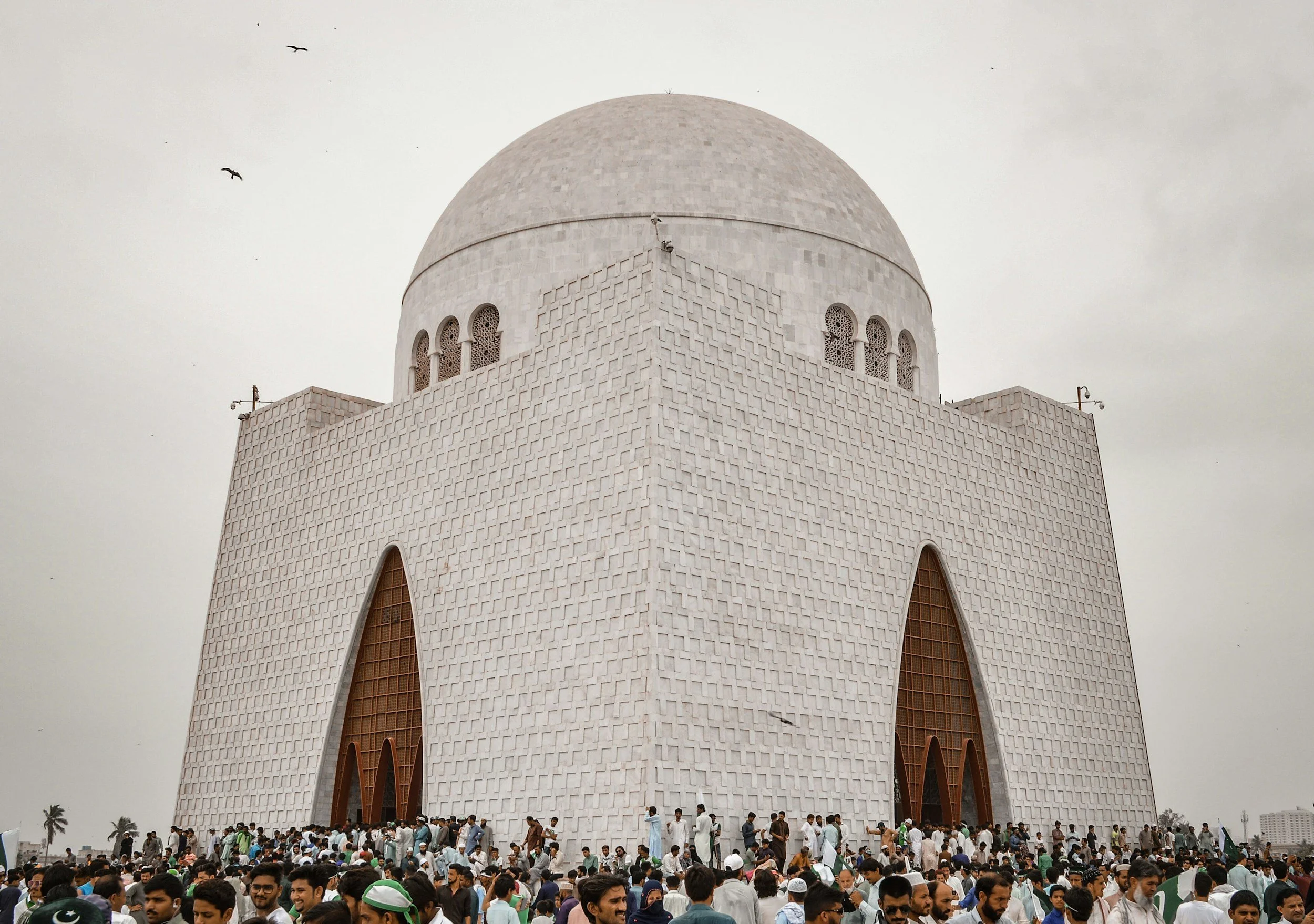 Large white mosque with a prominent dome and arched doorways, crowd of people gathered at the base, some waving Pakistani flags, under a cloudy sky.