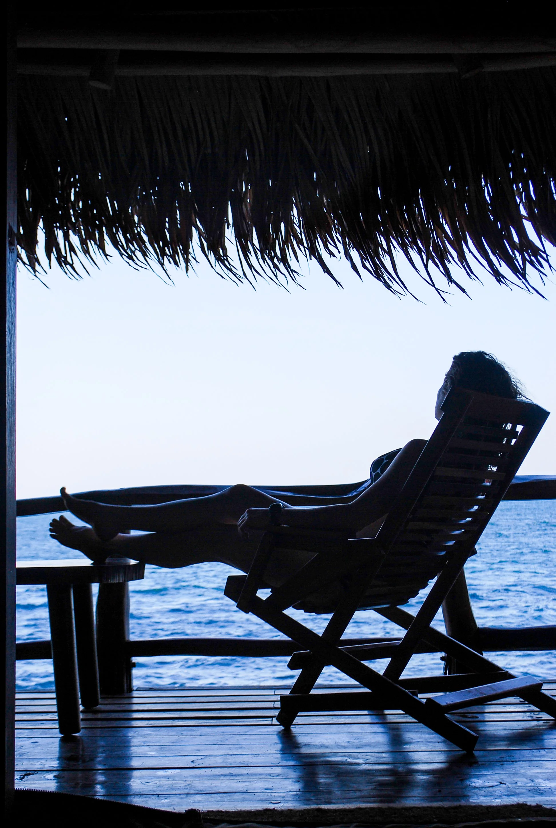 Person relaxing on a lounge chair on a thatched-roof deck overlooking the ocean.