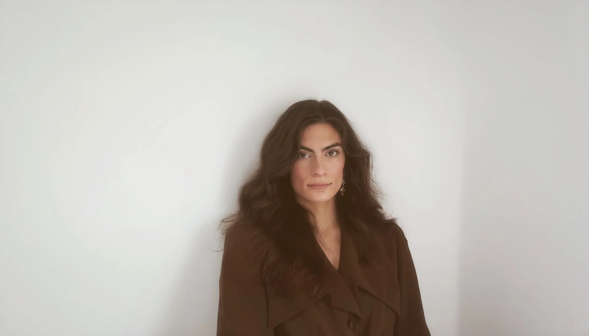 A woman with wavy dark brown hair and light makeup, wearing a brown blazer, standing against a plain white wall.