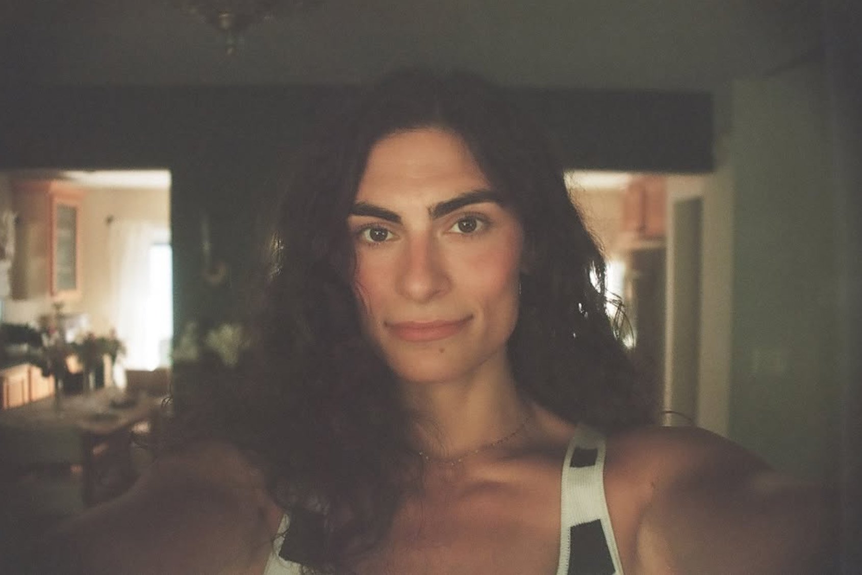 A woman taking a selfie of her reflection in a mirror inside a home, with a kitchen and dining area visible in the background.