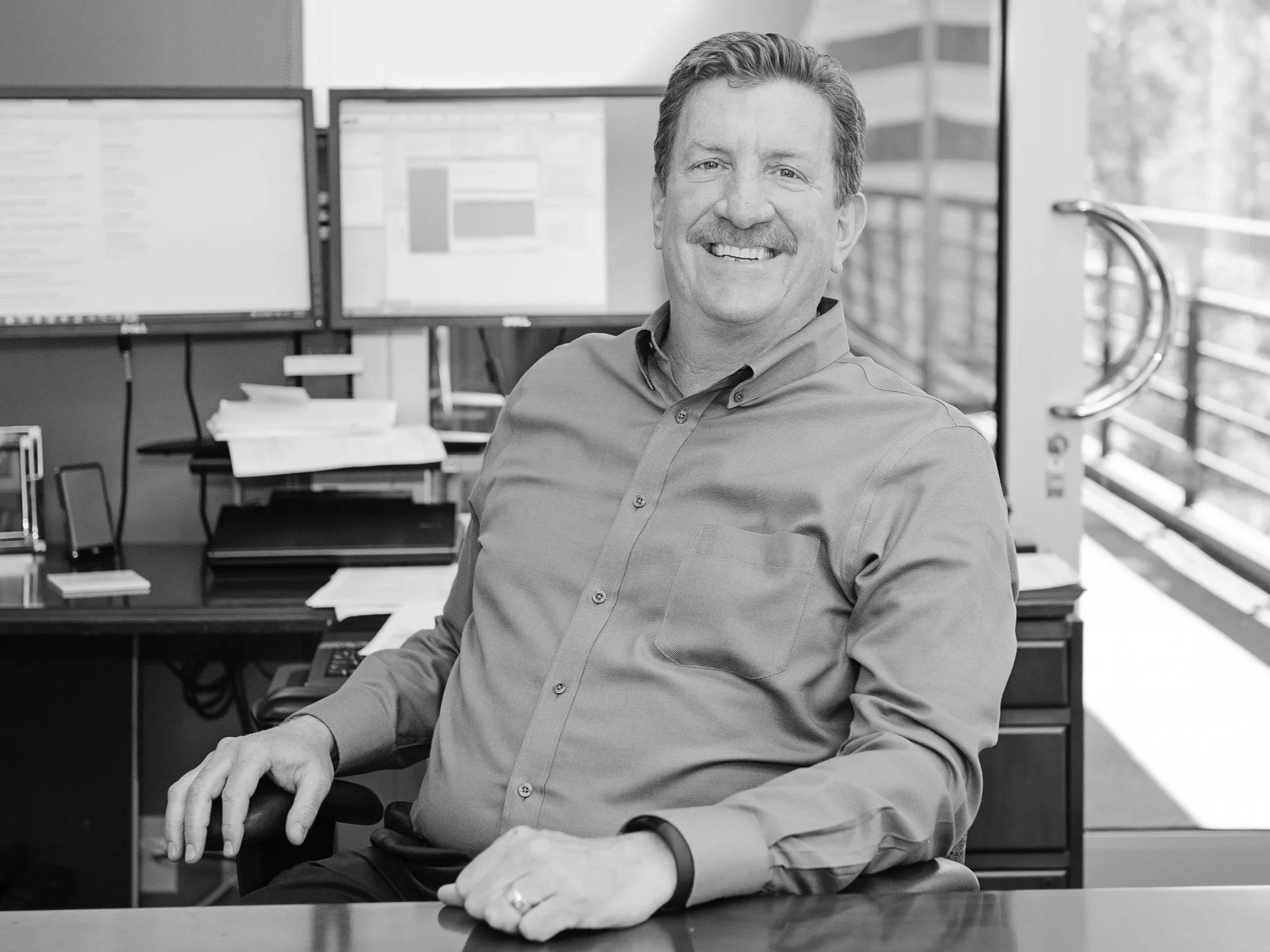 Peter Trumbo, a smiling man with a mustache sitting behind a desk with two screens behind him