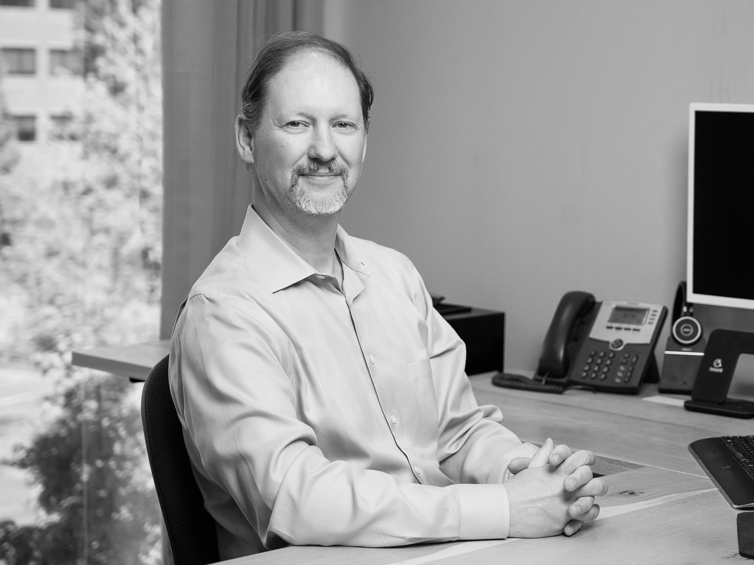 Mike McDougall, a smiling man sitting at his desk with clasped hands