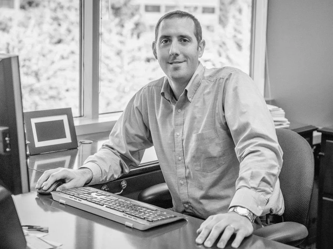 Adam Wilkie, a smiling man sitting at his computer with one hand on the mouse