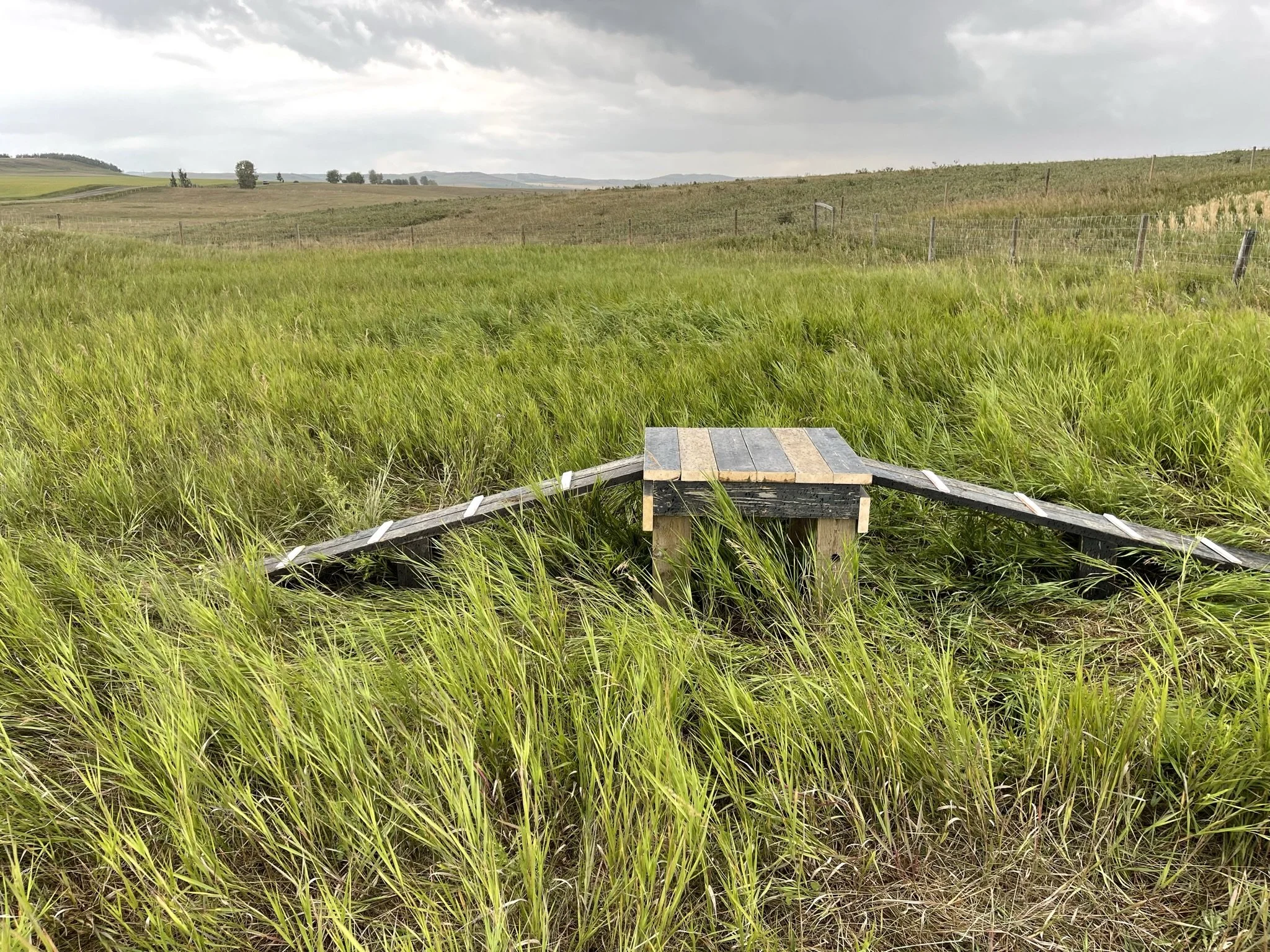 A scenic view of open farmland with green and brown fields, a barbed wire fence, and a cloudy sky overhead.