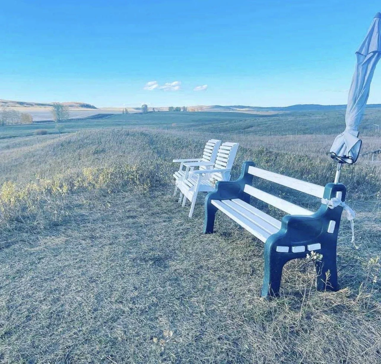 Empty benches and a closed umbrella on a grassy field with a vast open landscape and blue sky in the background.