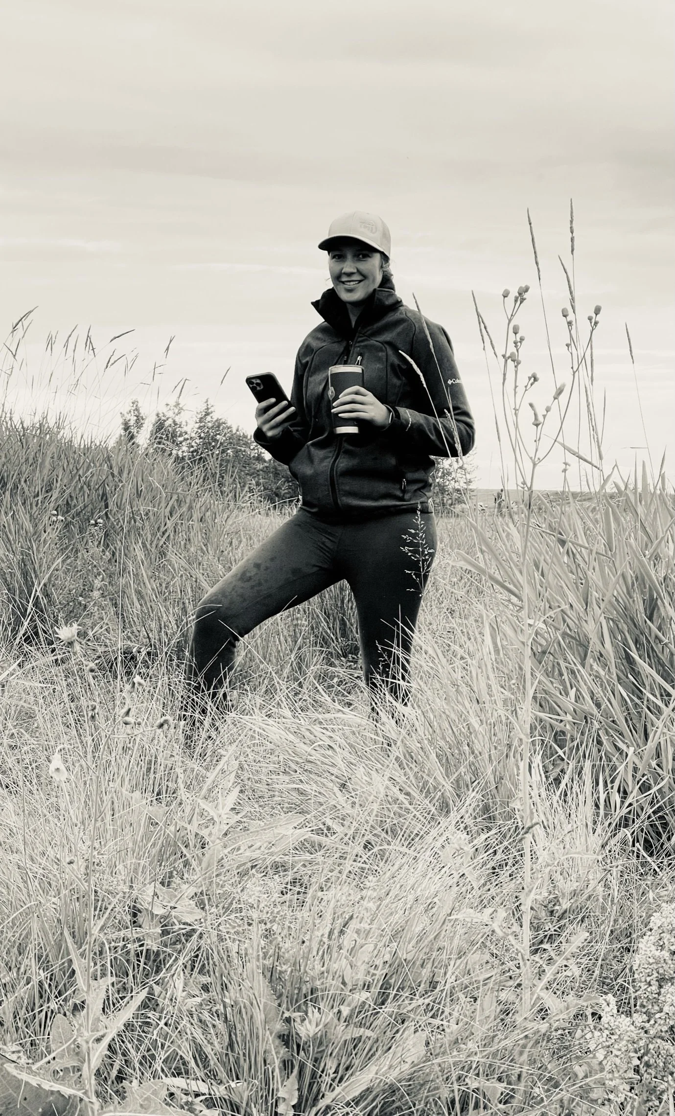 A woman standing in a field of tall grass, smiling, wearing a cap, jacket, and holding a phone and a tumbler.