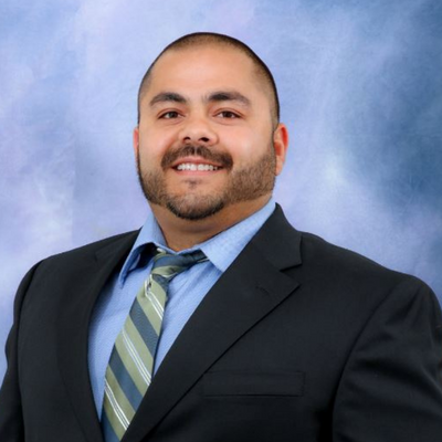 A man with a beard and short hair wearing a dark suit, light blue shirt, and striped tie, smiling against a blue background.