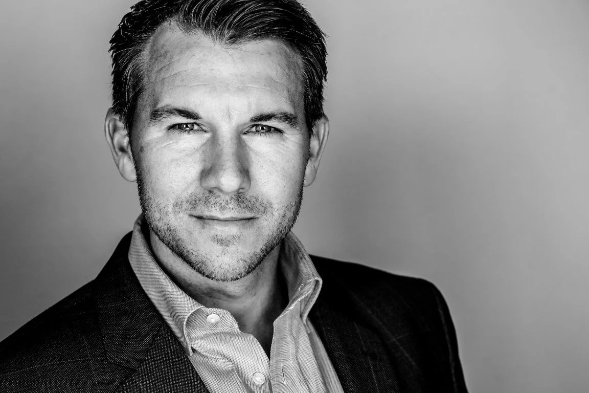 Black and white headshot of a man with short dark hair, wearing a collared shirt and blazer, looking at the camera with a serious expression.