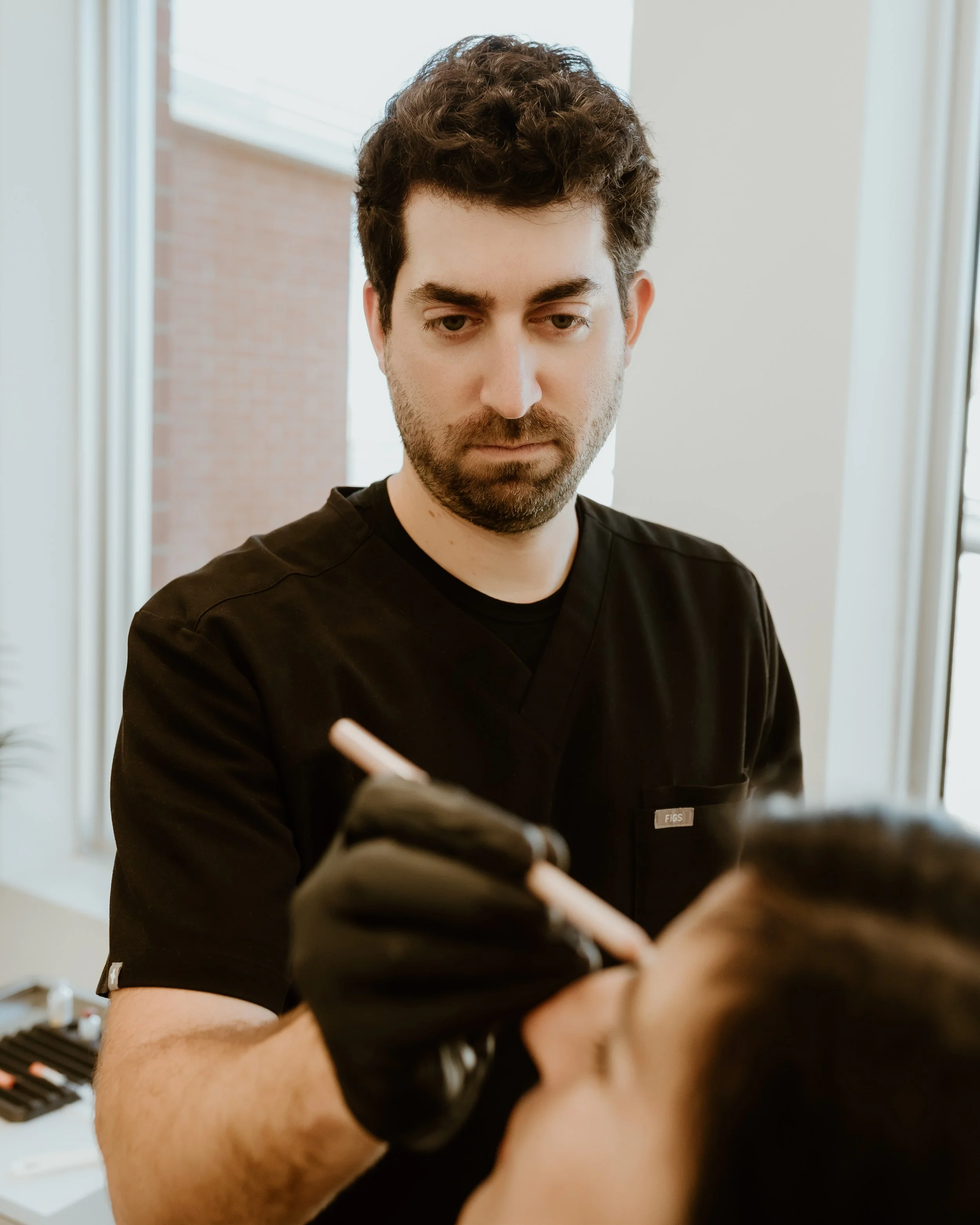 Un professionnel de la santé, portant un uniforme noir, réalise une injection ou un traitement sur un patient allongé, dans un environnement intérieur lumineux.