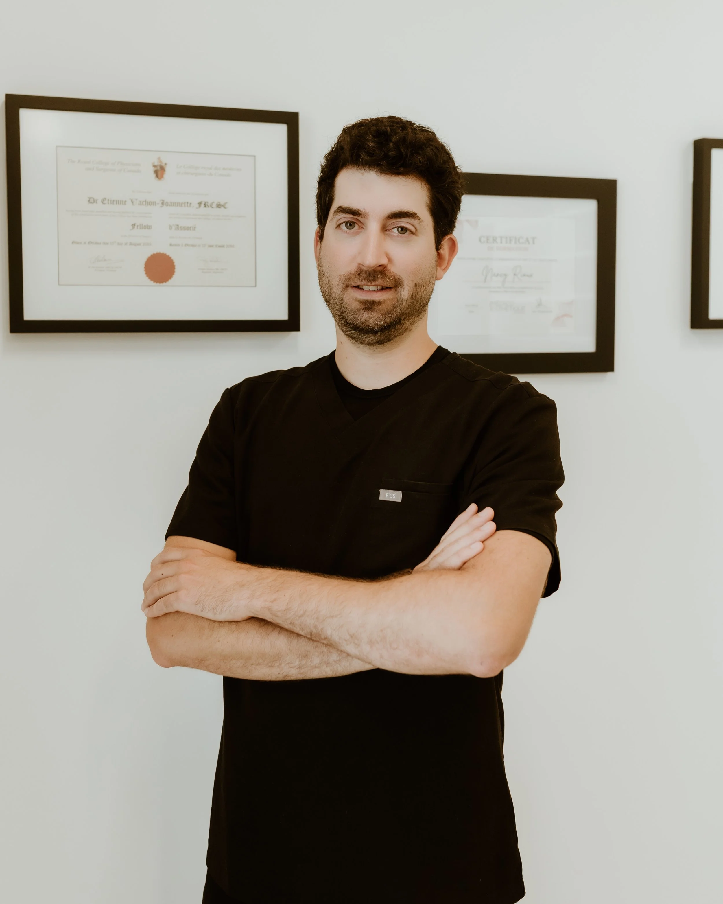Homme en blouse noire avec bras croisés, debout devant un mur blanc avec des diplômes encadrés.