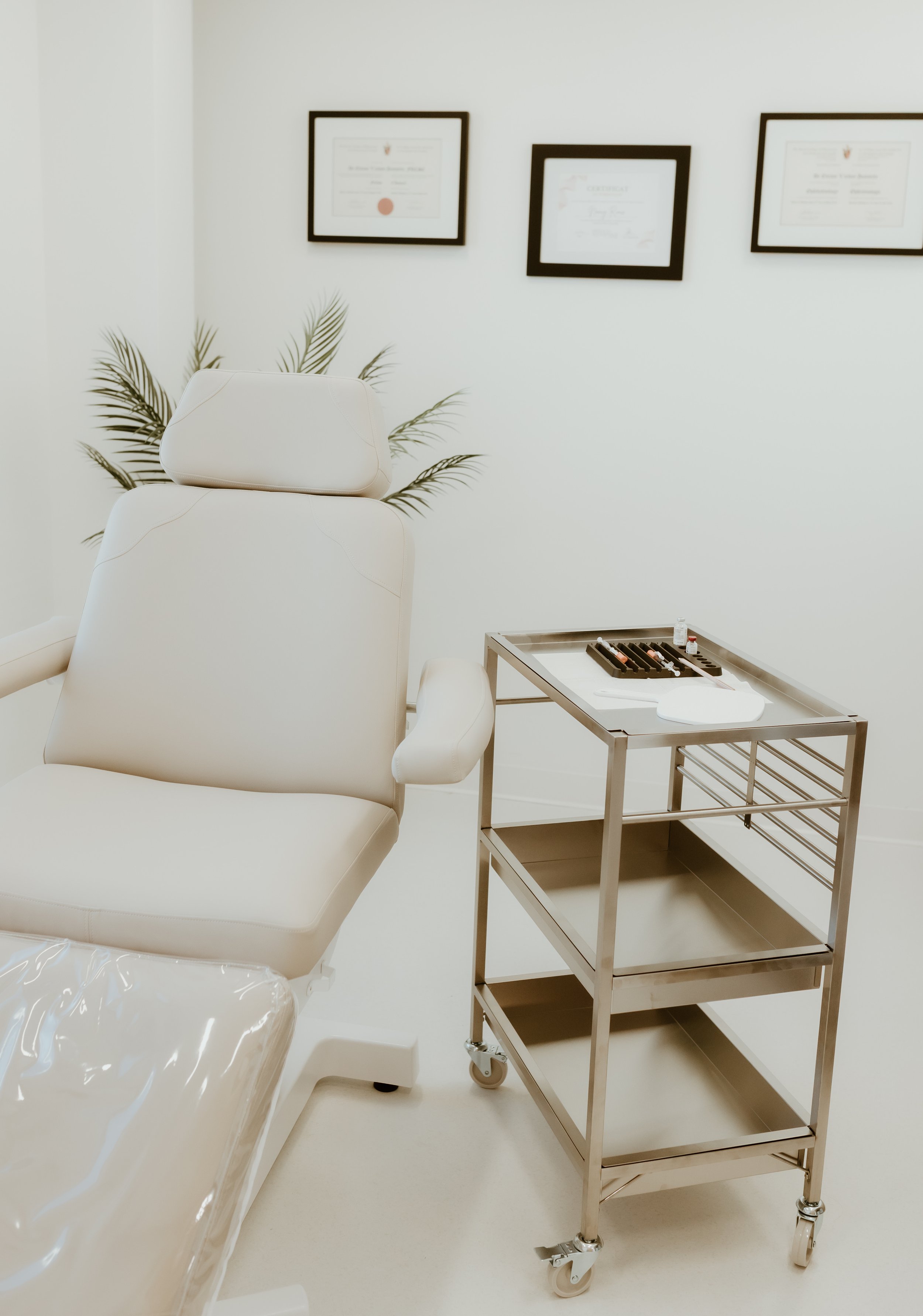 Une salle de clinique ou de dispensaire avec un fauteuil blanc, un chariot en métal avec des instruments médicaux et des diplômes encadrés au mur.