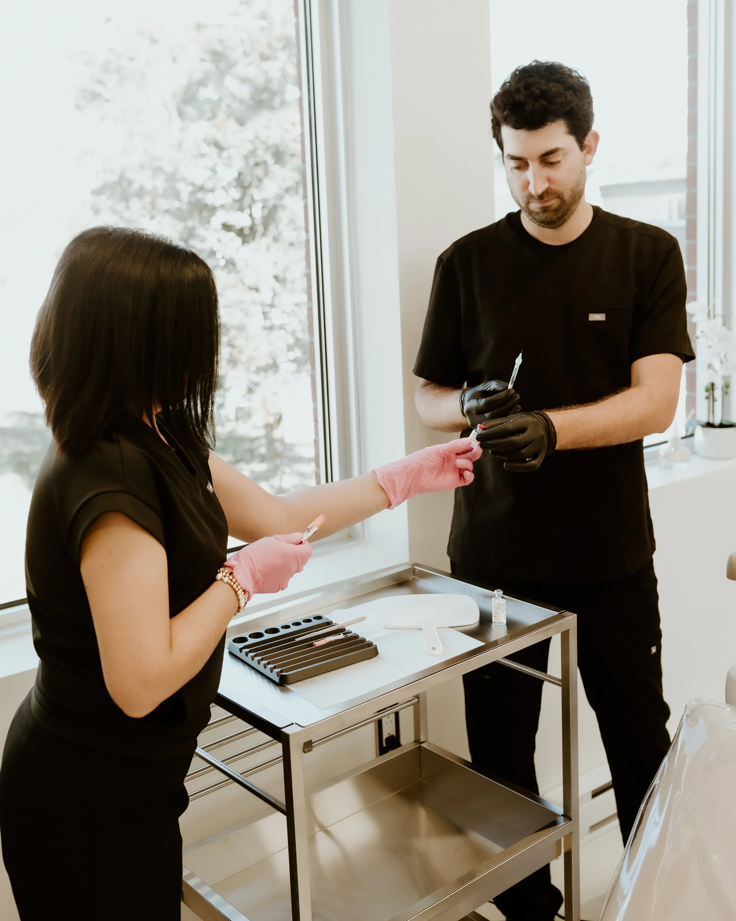 Un homme et une femme dans une clinique ou un hôpital, en train de faire une injection, portant des gants noirs et roses respectivement, avec une table contenant des seringues, un miroir, une petite bouteille et des tubes de test.