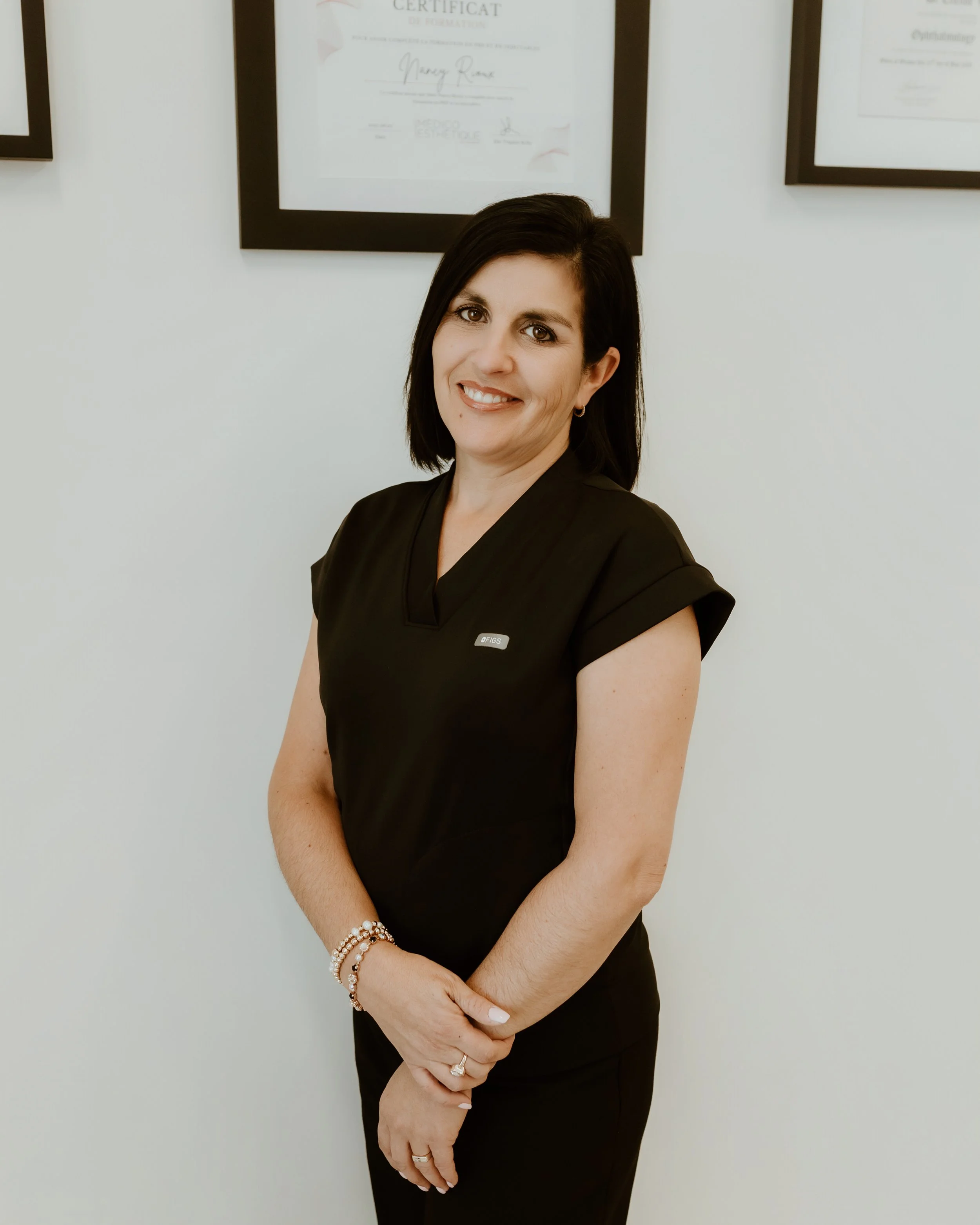 Une femme souriante avec des cheveux noirs, portant une tenue noire, se tenant devant un mur avec des diplômes encadrés.