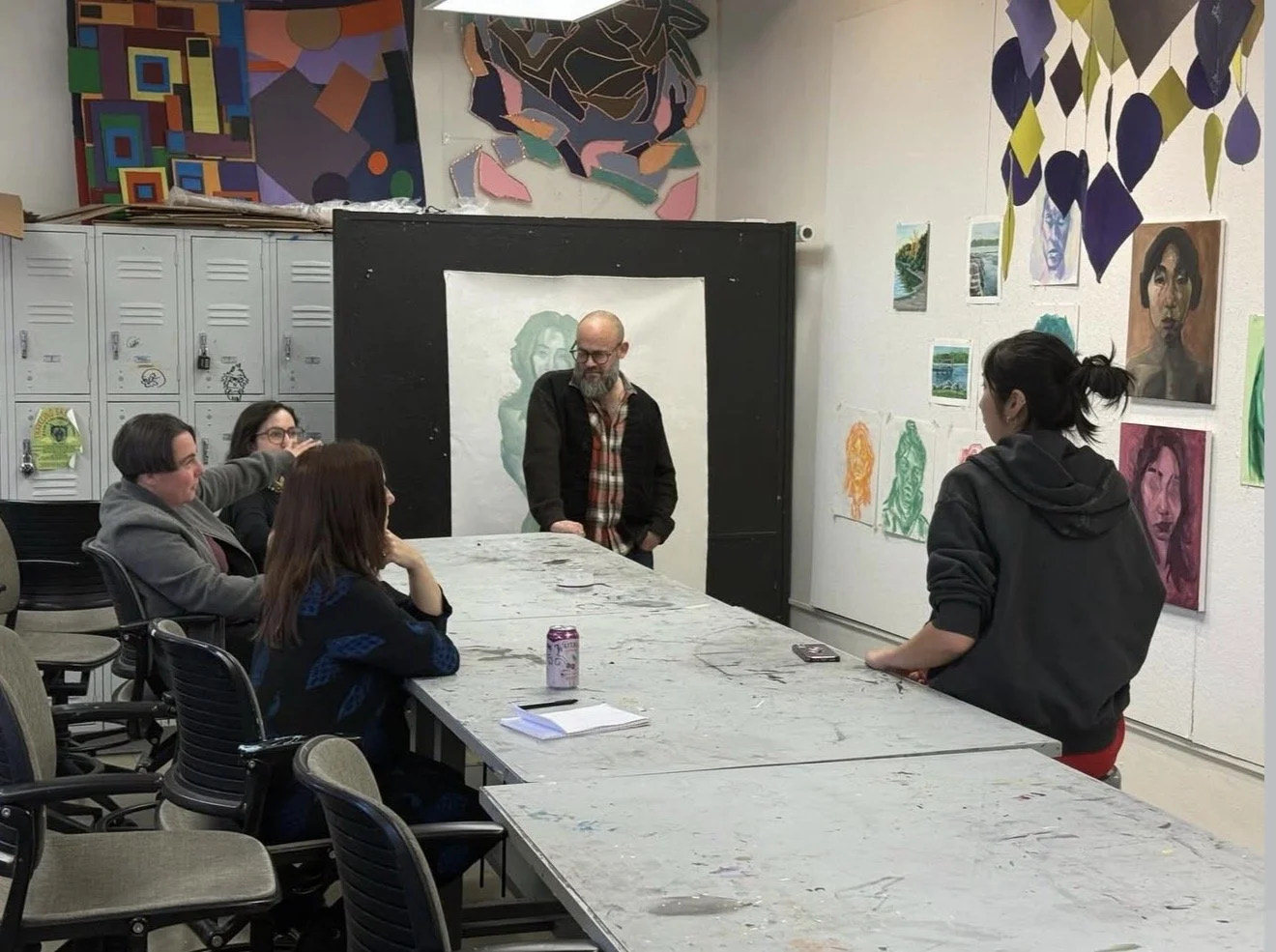 Art classroom with six people around a large table, four sitting and two standing, in a room decorated with colorful paintings and drawings, including a blackboard with a portrait of a woman.