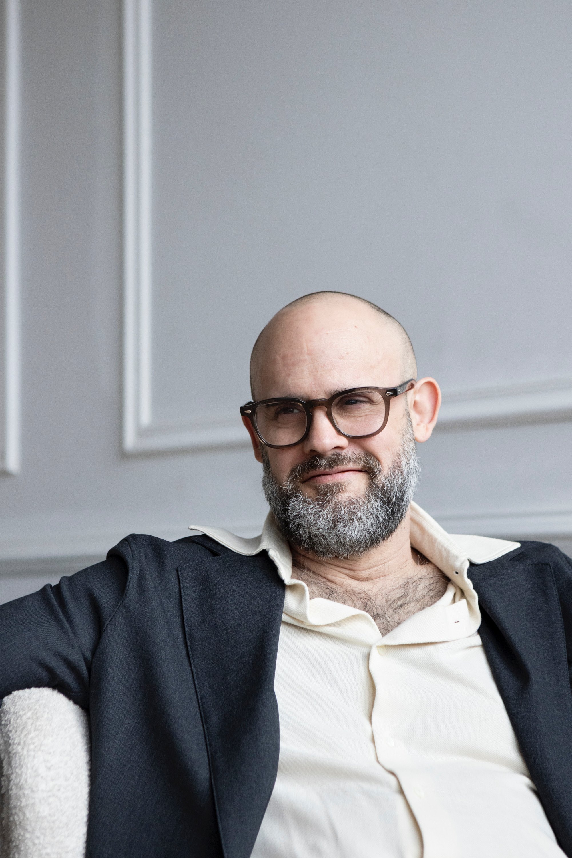 A man with glasses and a beard, dressed in a white shirt and dark blazer, sitting on a beige chair against a wall with white trim.